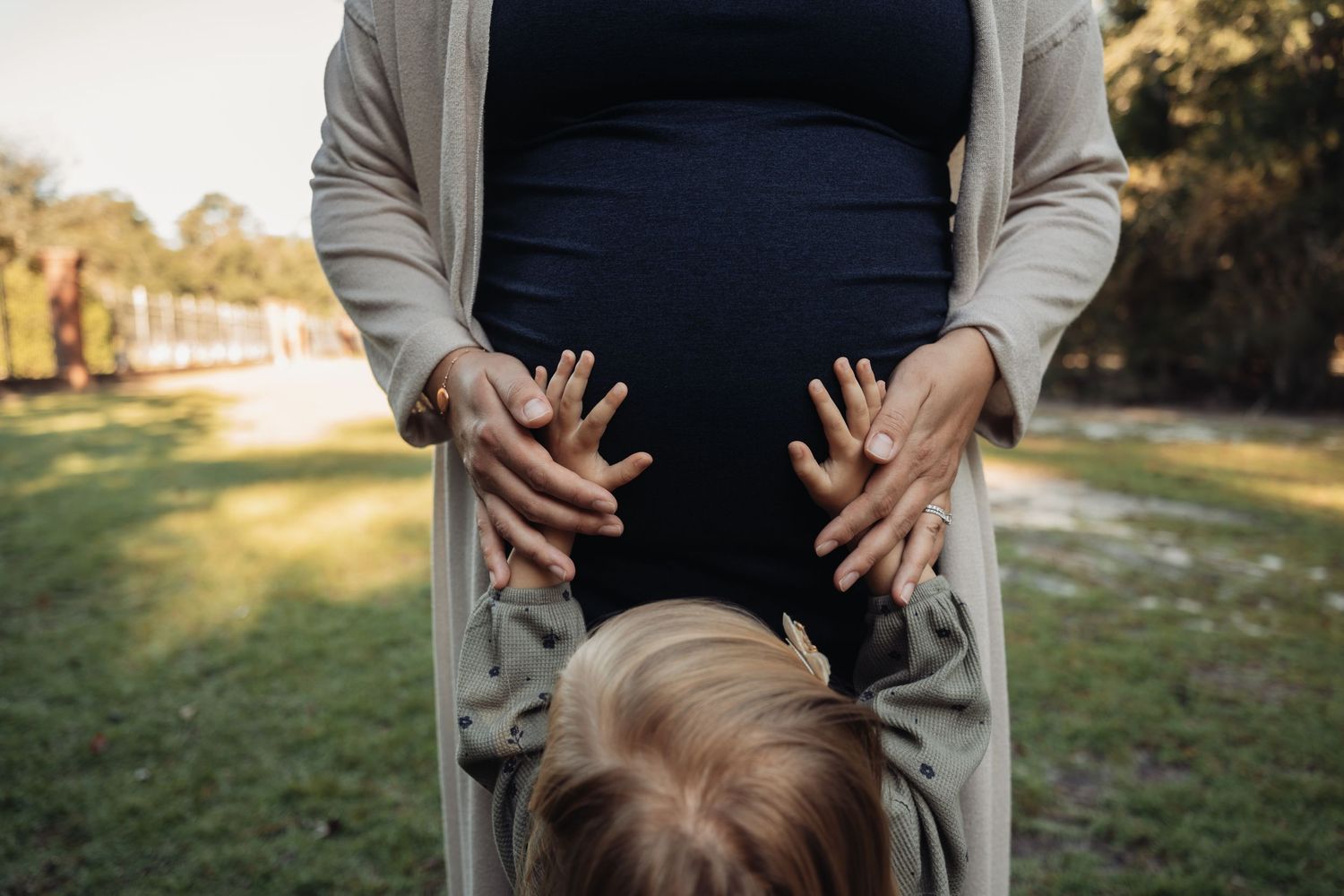 Pregnant woman in black dress with young child resting head on baby bump outdoors in park setting.