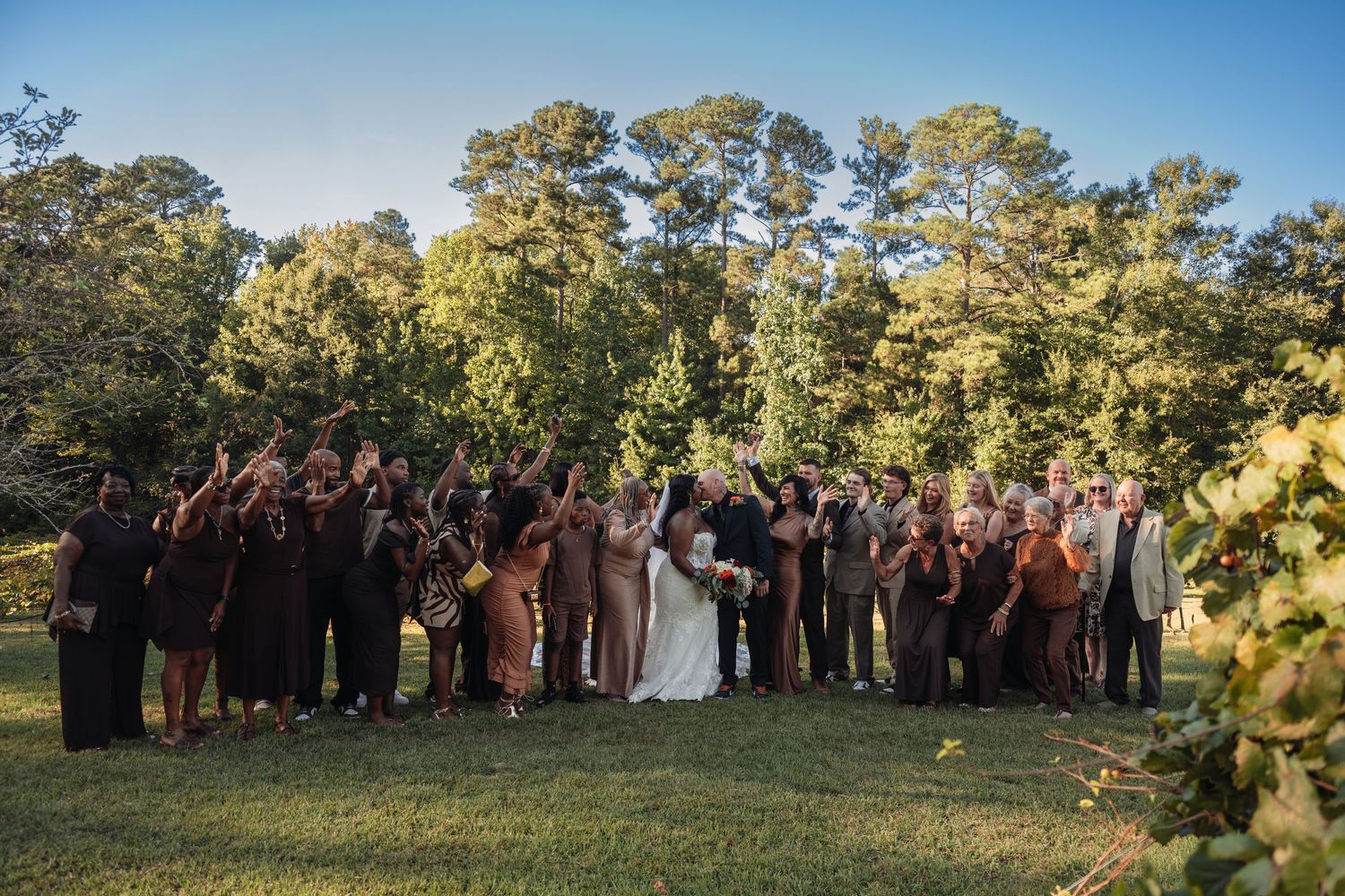 Large wedding party group photo on a sunny lawn, all cheering with hands raised behind the couple.