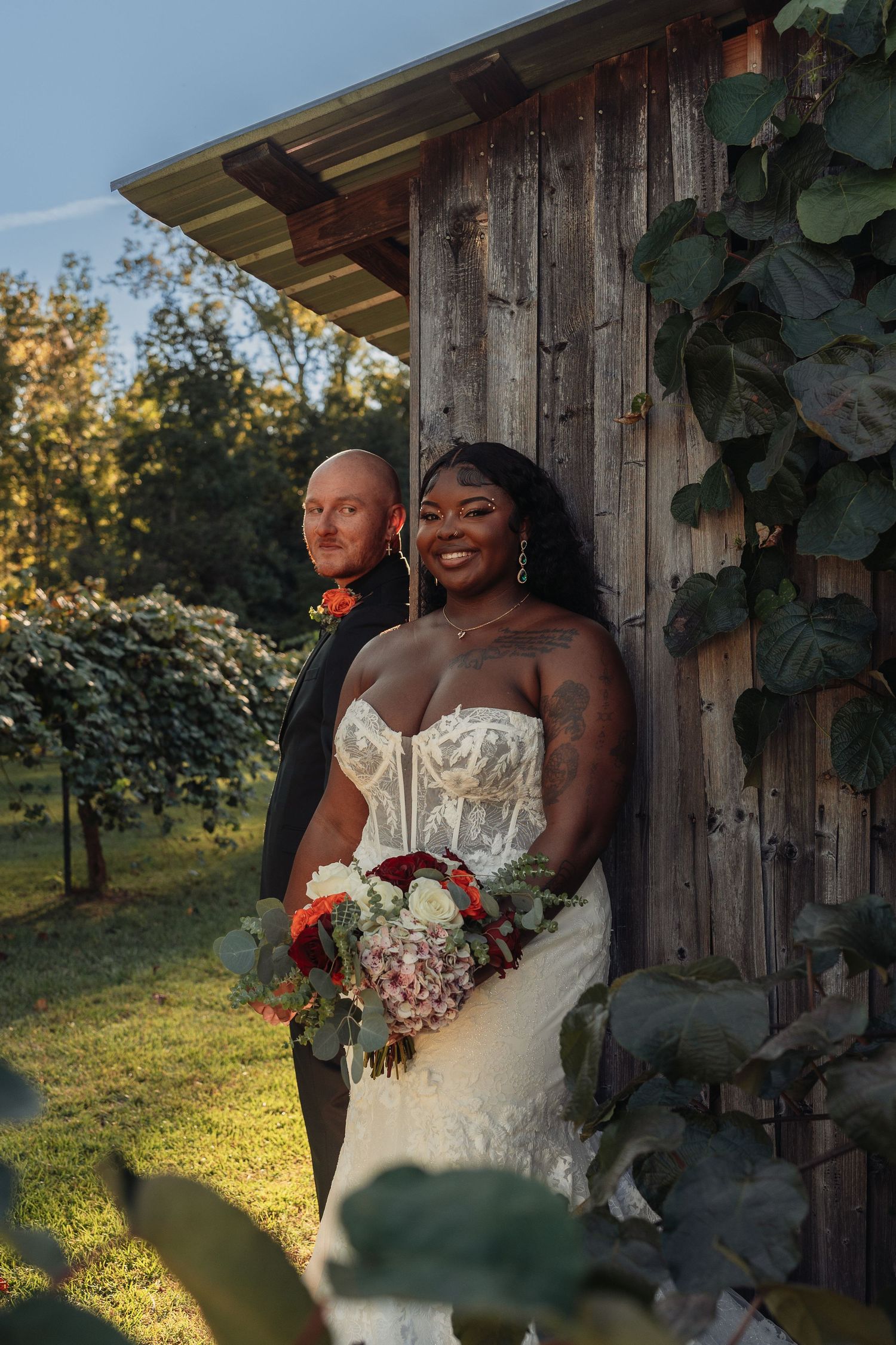 Bride and groom posing by rustic wooden barn wall covered in green ivy vines.