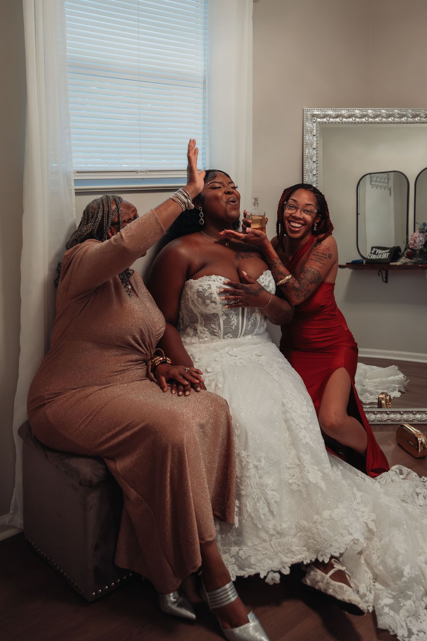 Bride laughs joyfully with two friends in red and beige dresses during wedding preparations indoors.
