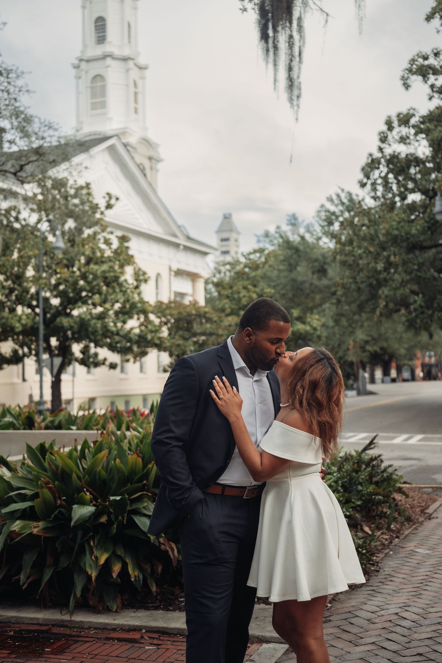 Couple sharing a kiss on Savannah street with historic white church steeple visible in background.