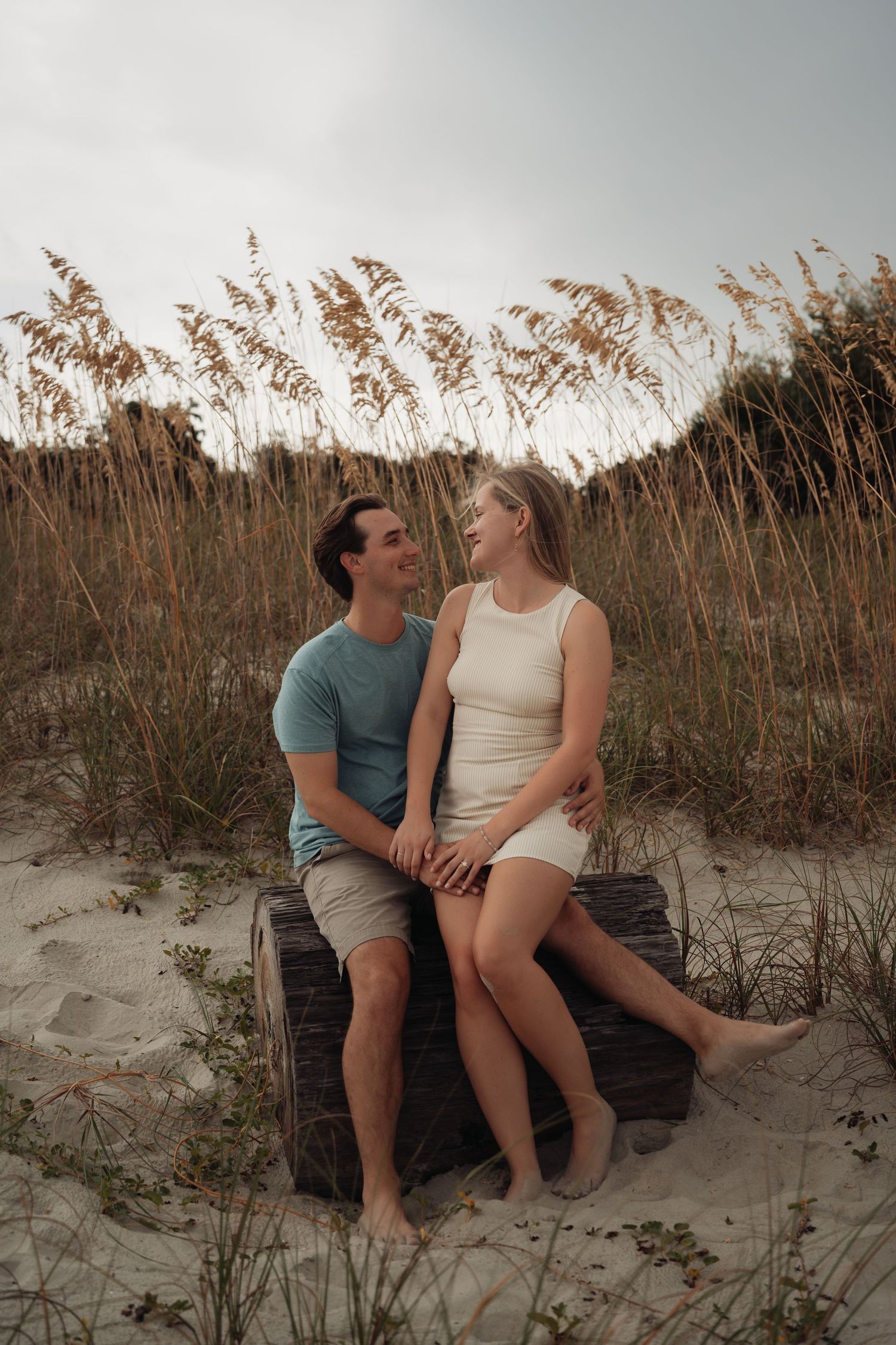A couple sits together on a wooden log in sandy beach dunes, smiling at each other with sea oats in the background.
