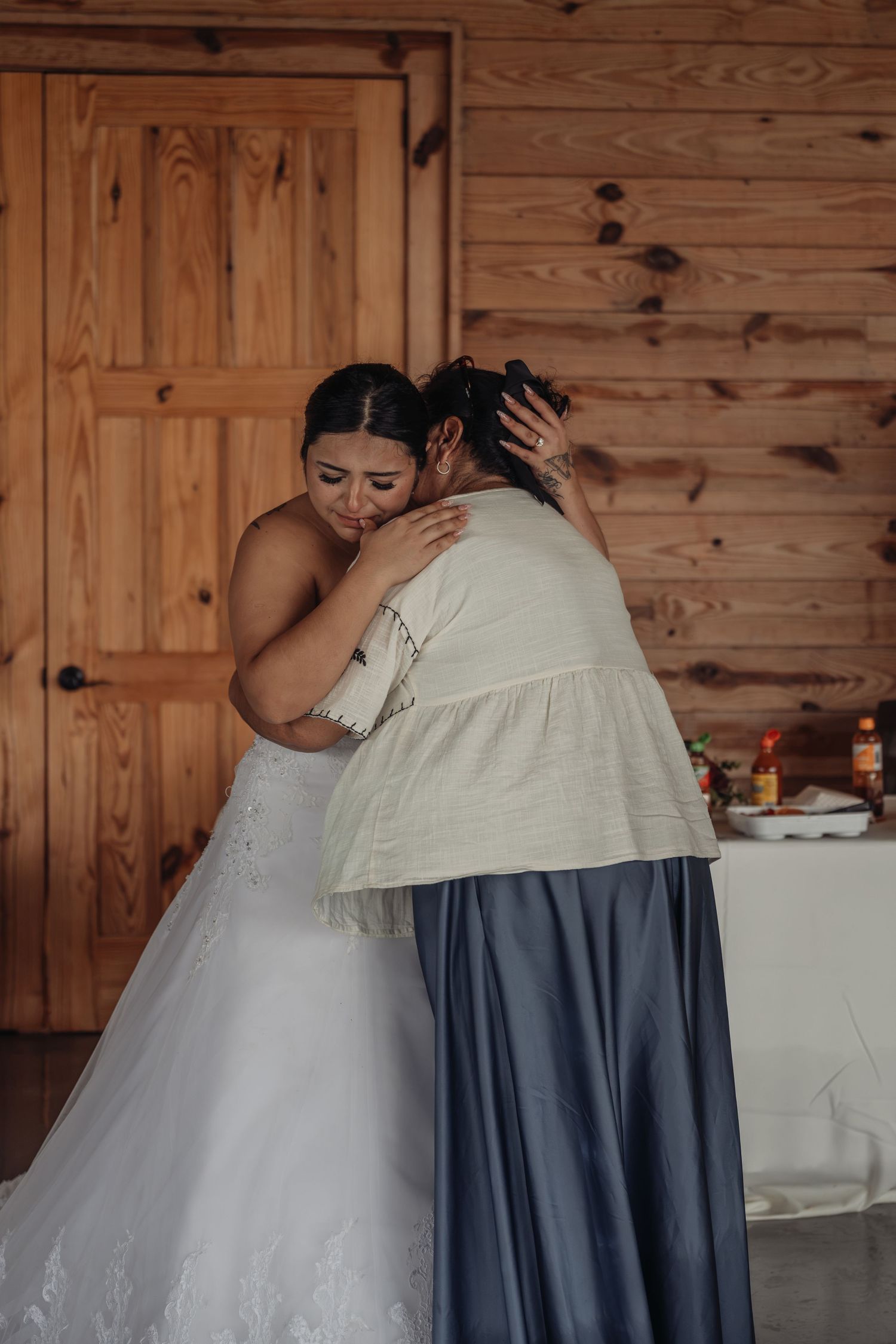 Bride in white wedding dress sharing an emotional hug with a guest inside a rustic wooden cabin venue.