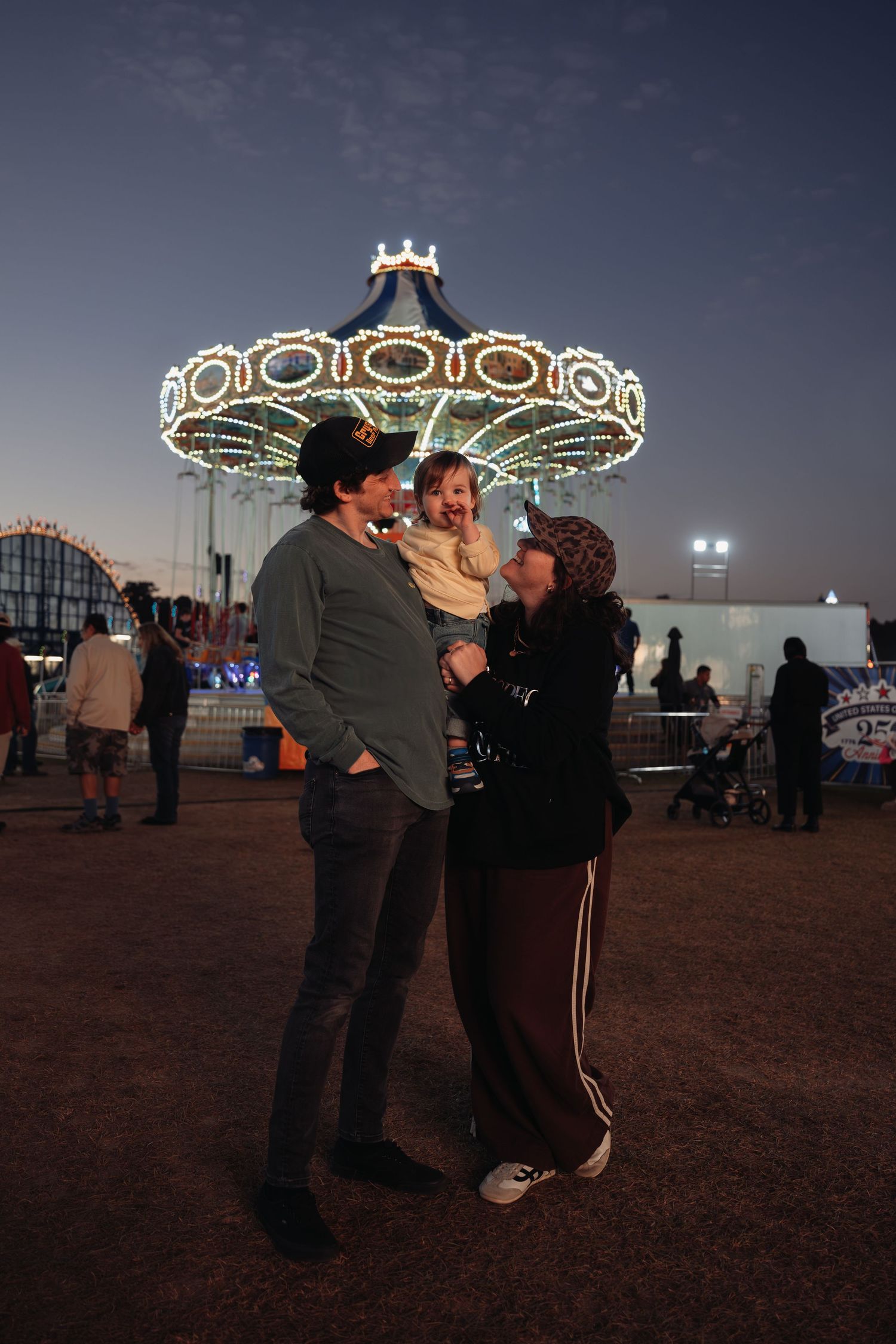 A couple holds a smiling toddler at a fairground at dusk, with a brightly lit swing carousel glowing in the evening sky behind them.