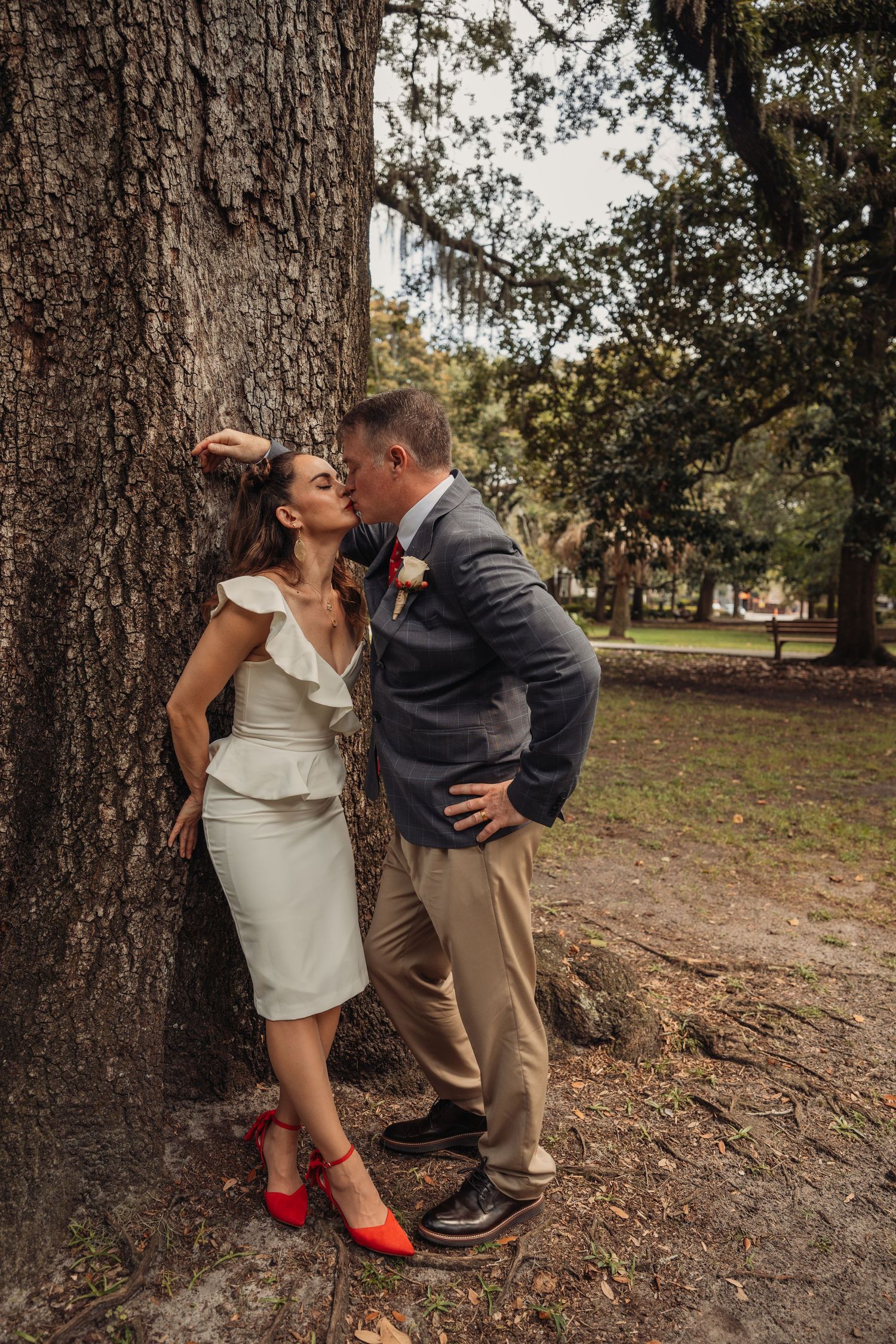A groom kisses his bride as she leans against a large oak tree in a scenic outdoor park setting.