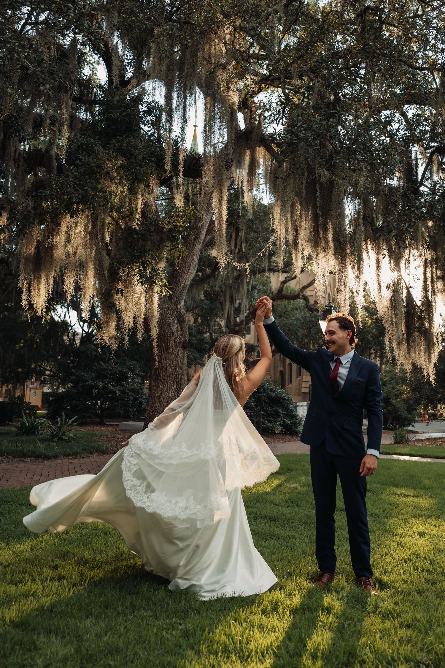 Groom twirls bride in flowing white gown under Spanish moss-draped oak trees at golden hour.