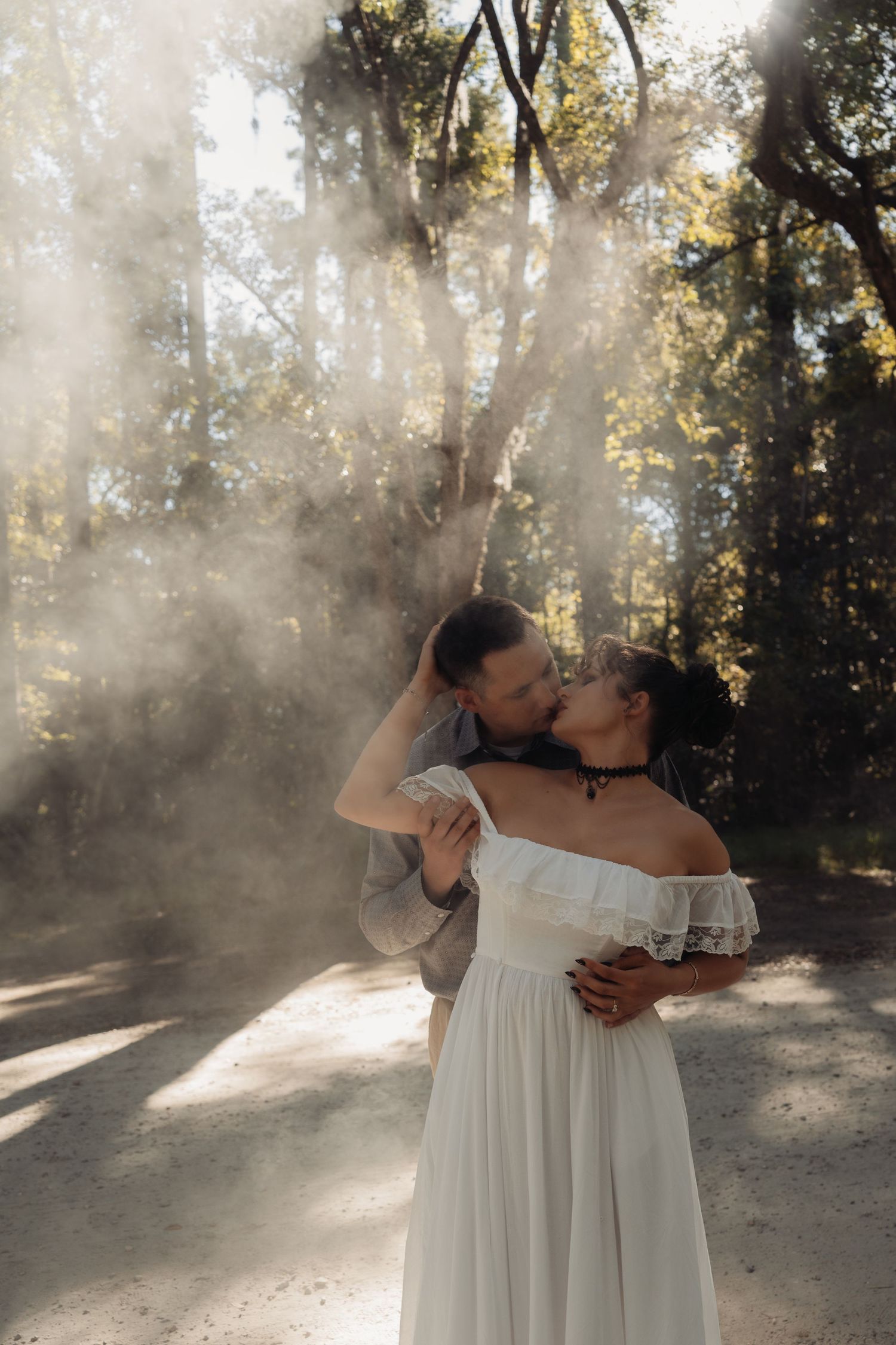 Couple sharing a passionate kiss in a sunlit forest with dramatic light rays, woman in white off-shoulder bridal gown.