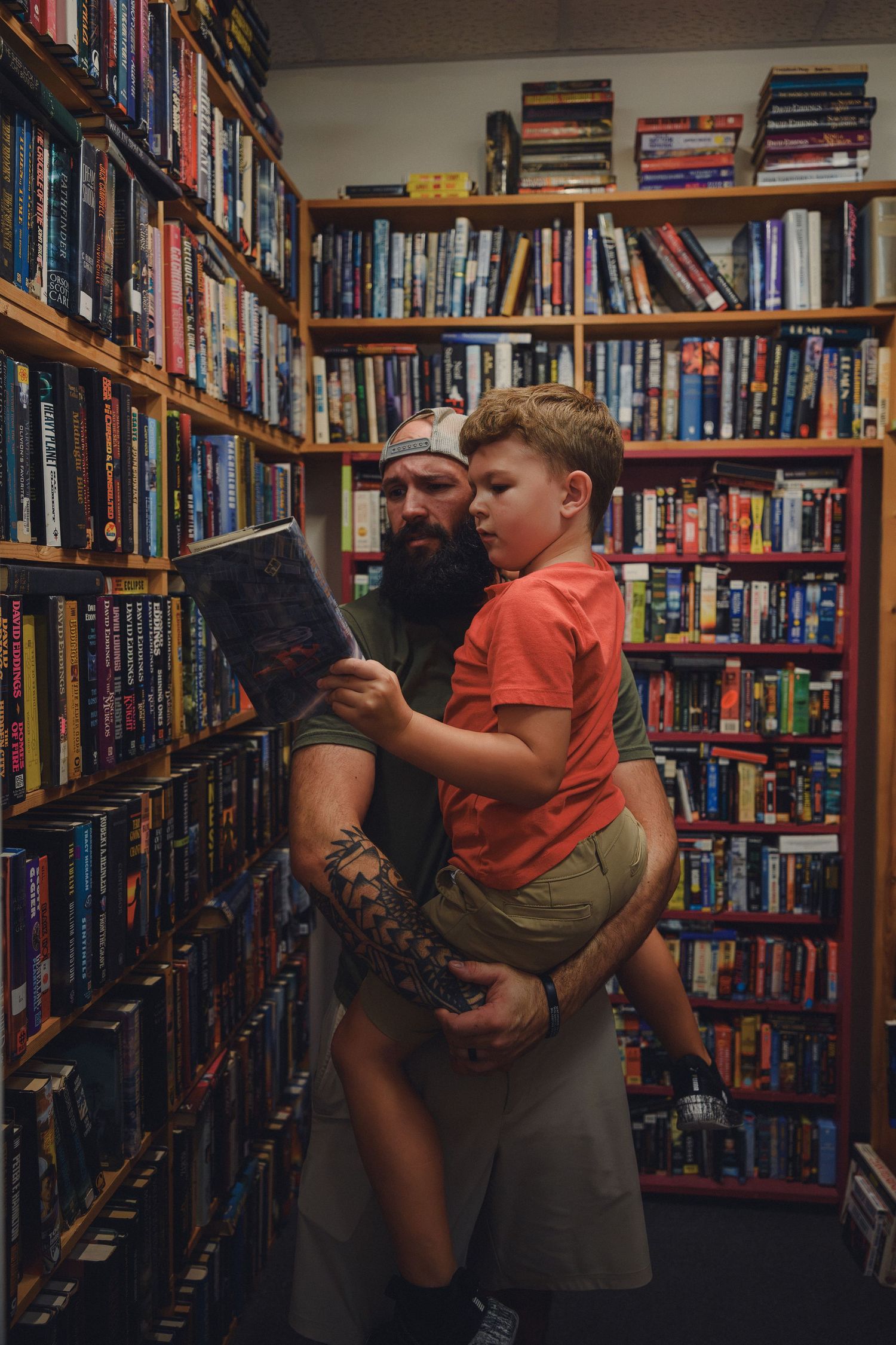 Tattooed father holds young son while browsing books in a cozy bookstore with colorful shelves.