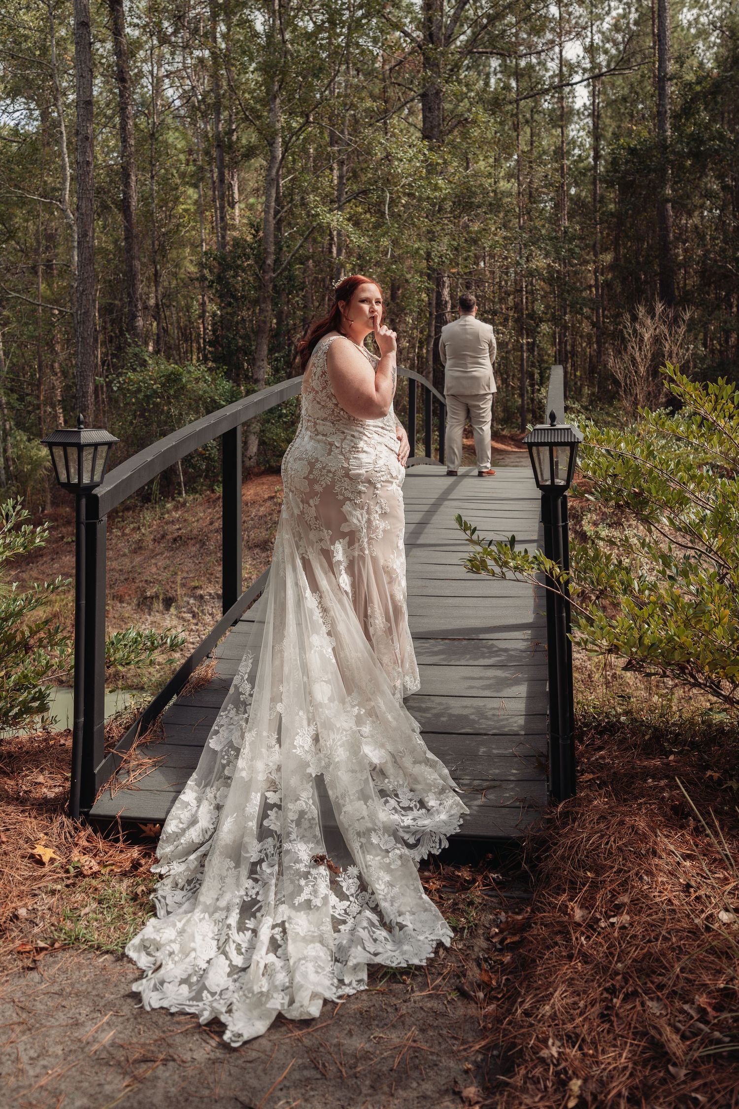 Bride in a lace wedding gown with a long train looks back over her shoulder on a wooden bridge as the groom waits in the forest.