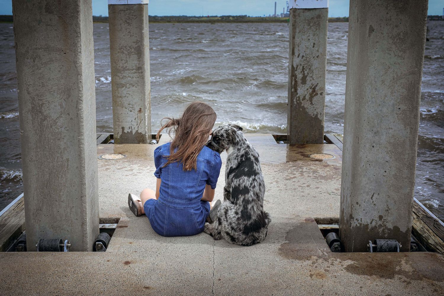 Girl and her dog sitting together facing the water, framed between wooden dock pillars, Jacksonville, FL