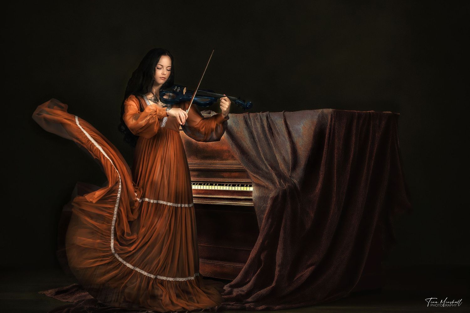 Woman in a flowing amber gown playing violin beside a vintage piano, moody dark editorial portrait, Jacksonville storytelling photography.