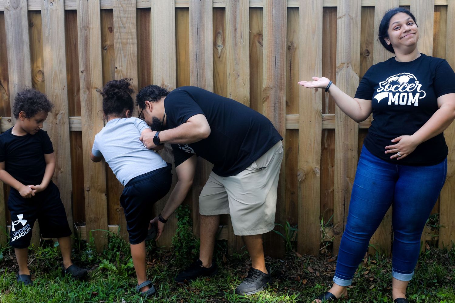 Family candid moment at a backyard fence, dad crouched with the kids while mom looks on laughing