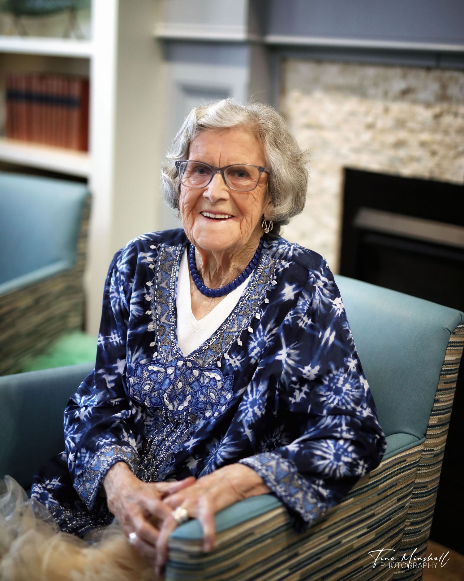 Older woman in a blue patterned top and glasses, seated portrait