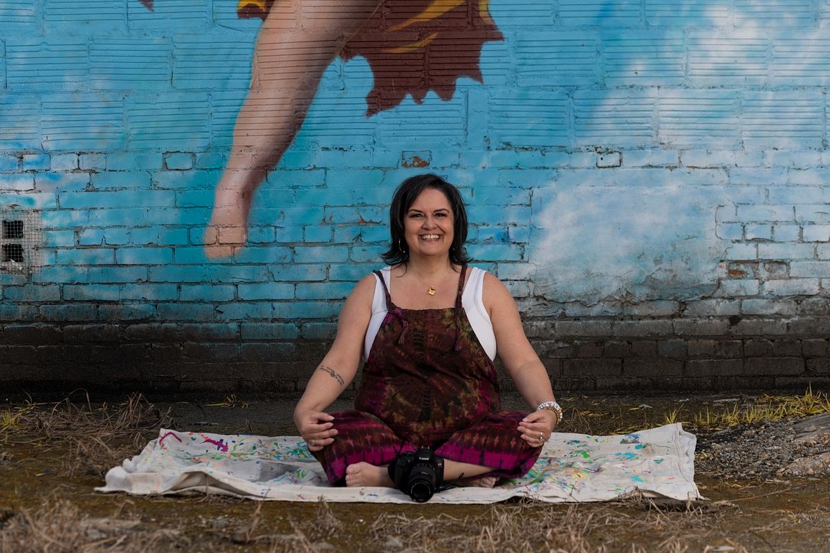 Tina Minshall, photographer, sitting cross-legged in front of a colorful mural in Jacksonville, FL, camera resting in her lap