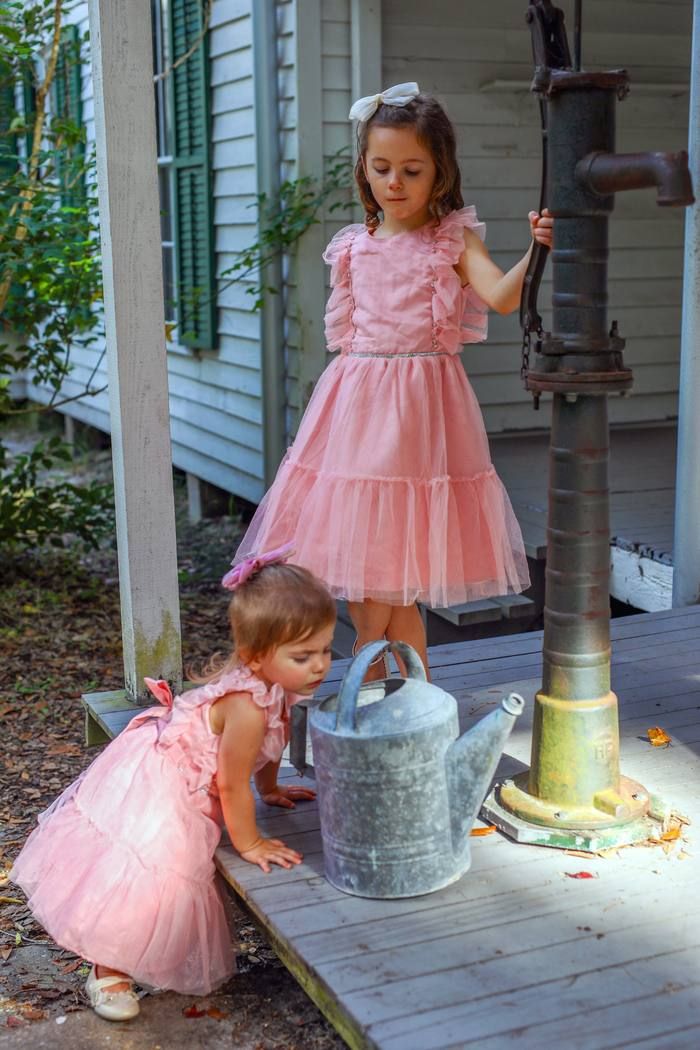 Two young sisters in pink dresses at an antique water pump on a porch, candid childhood moment