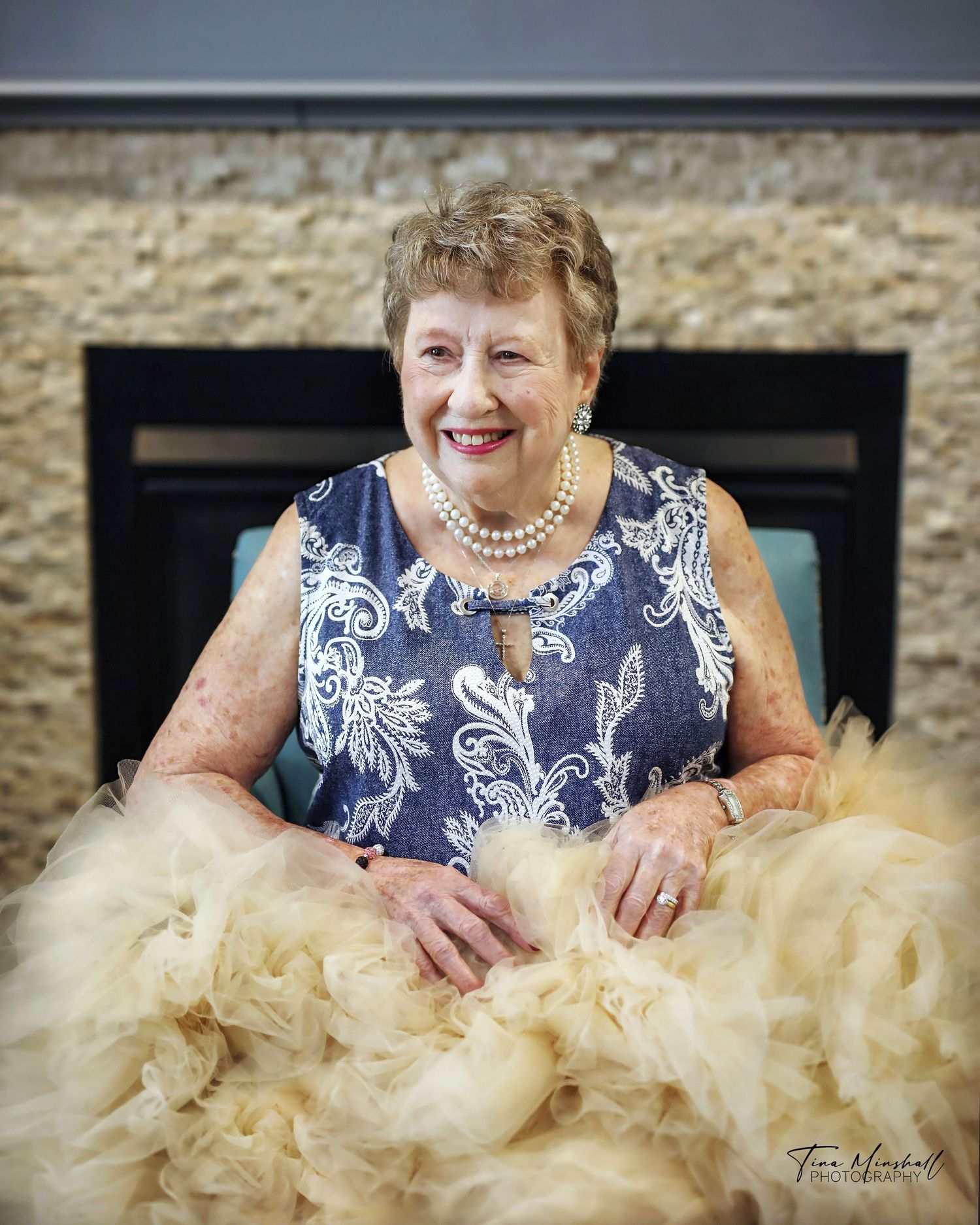 Older woman in a blue floral dress surrounded by a cream feather boa, warm portrait by the fireplace