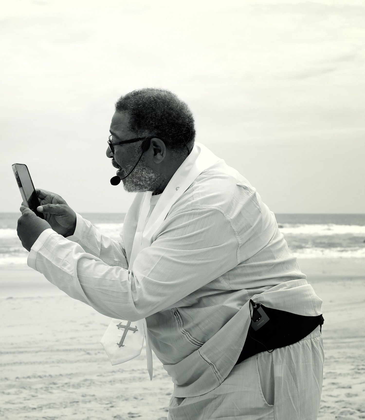 Man in white ceremonial robe photographing the ocean on his phone, black and white editorial portrait