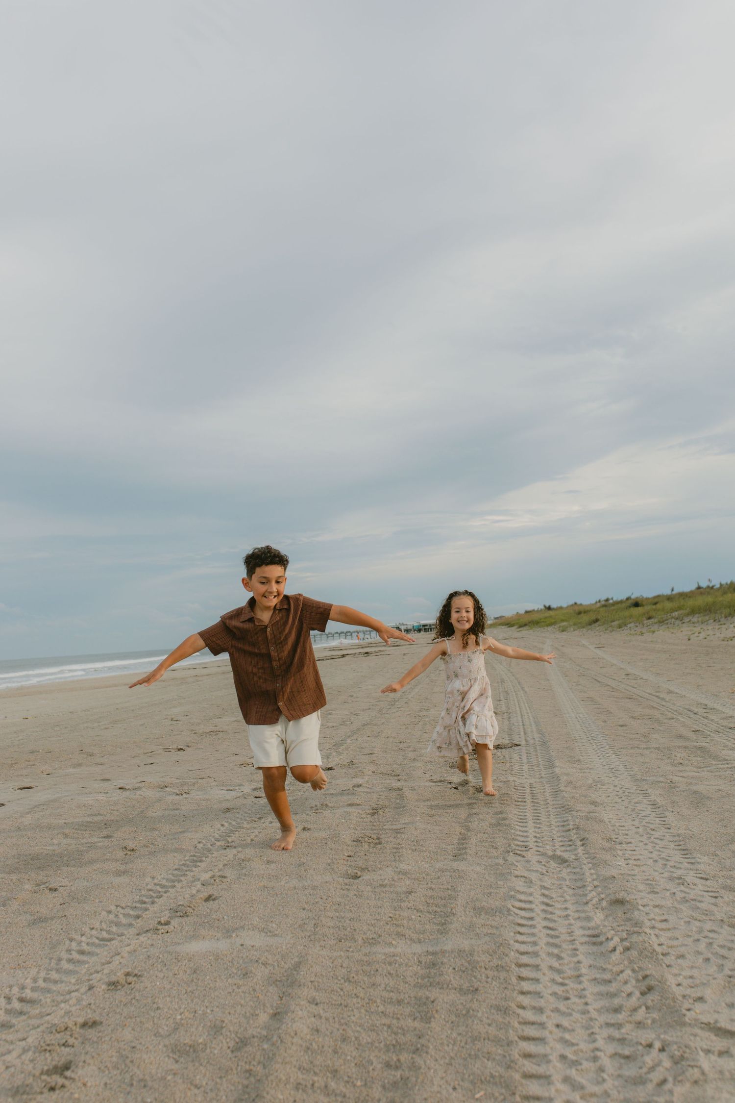 Two children running joyfully on a sandy beach with arms outstretched under a cloudy sky.