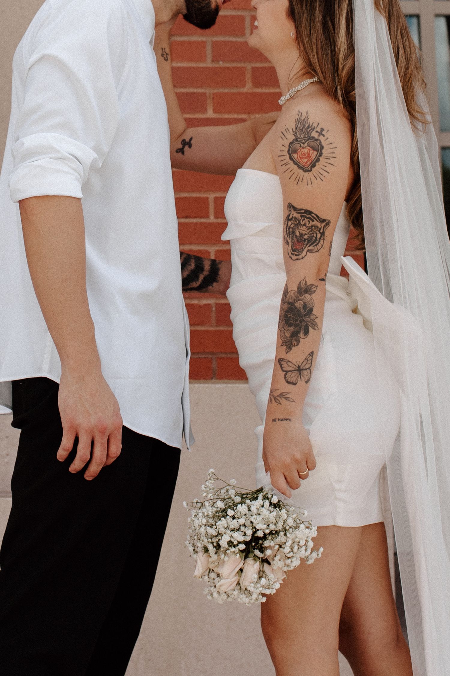Tattooed bride in short white dress holding baby's breath bouquet kissing groom near brick wall.