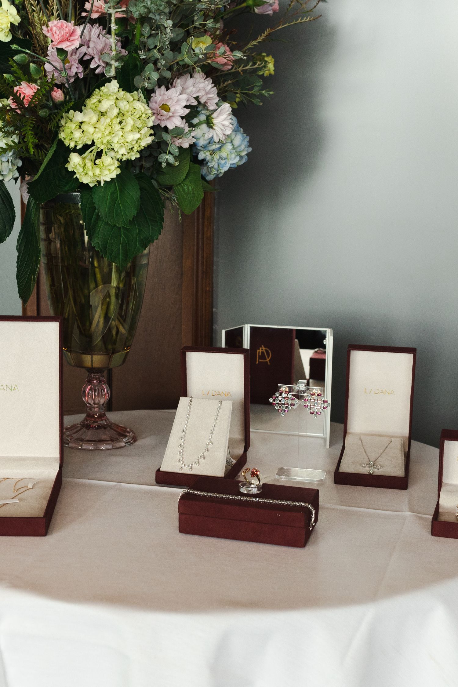 Elegant jewelry display with necklaces, earrings, and rings in burgundy boxes on a white table with floral arrangement.