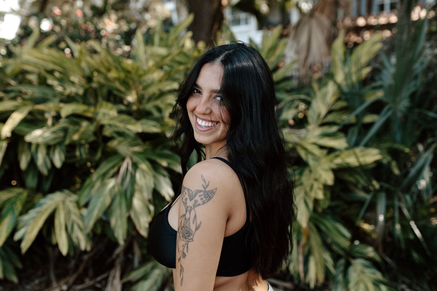 Smiling woman with shoulder tattoo posing in front of lush tropical green foliage.