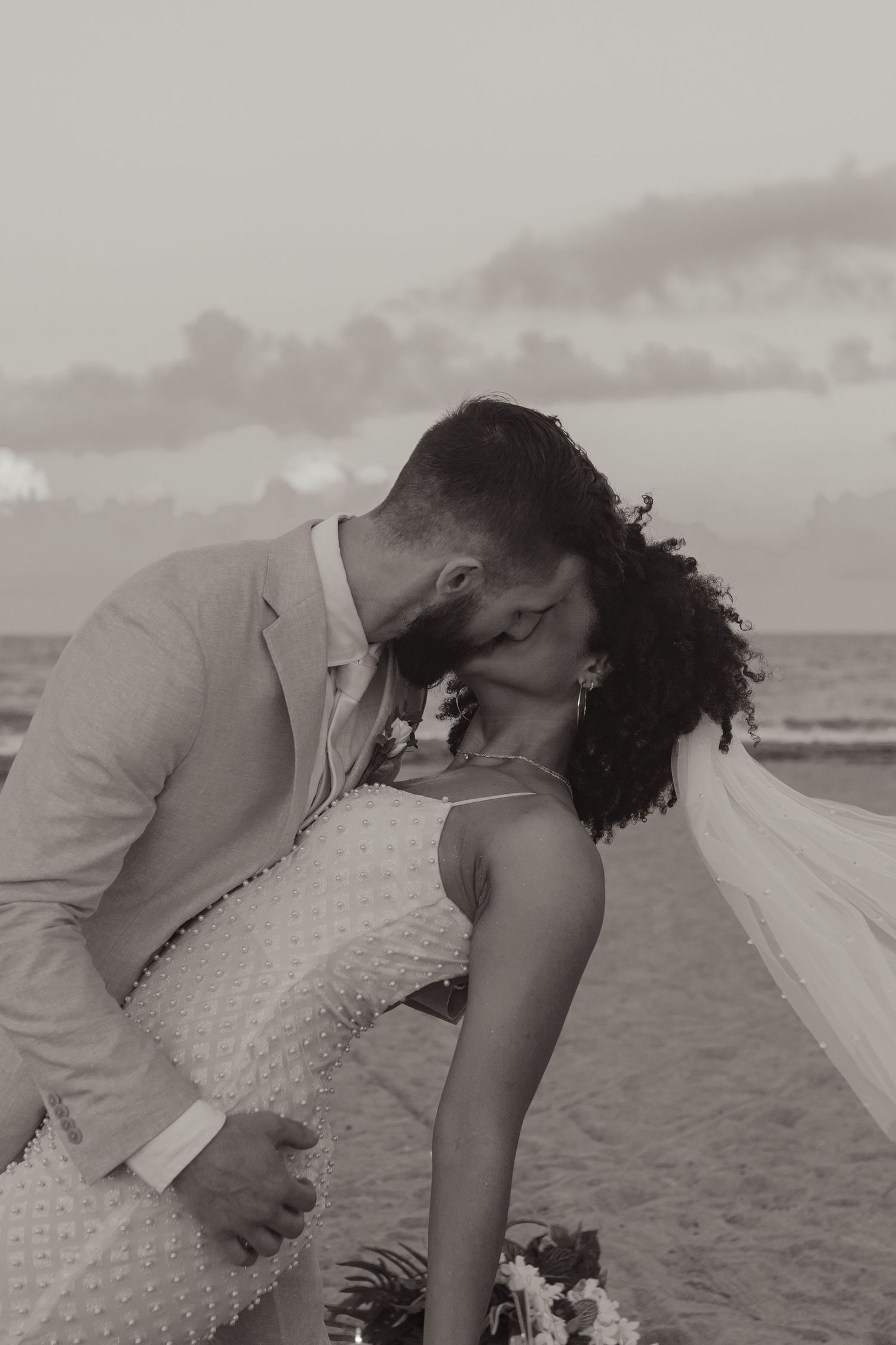 Bride and groom share an intimate kiss on the beach at sunset in a black and white photo.