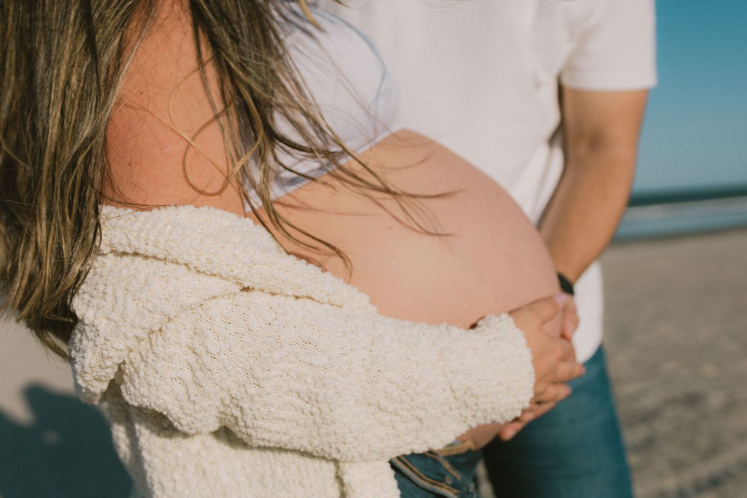 Pregnant woman in white knit cardigan holds baby bump on sunny beach with partner.