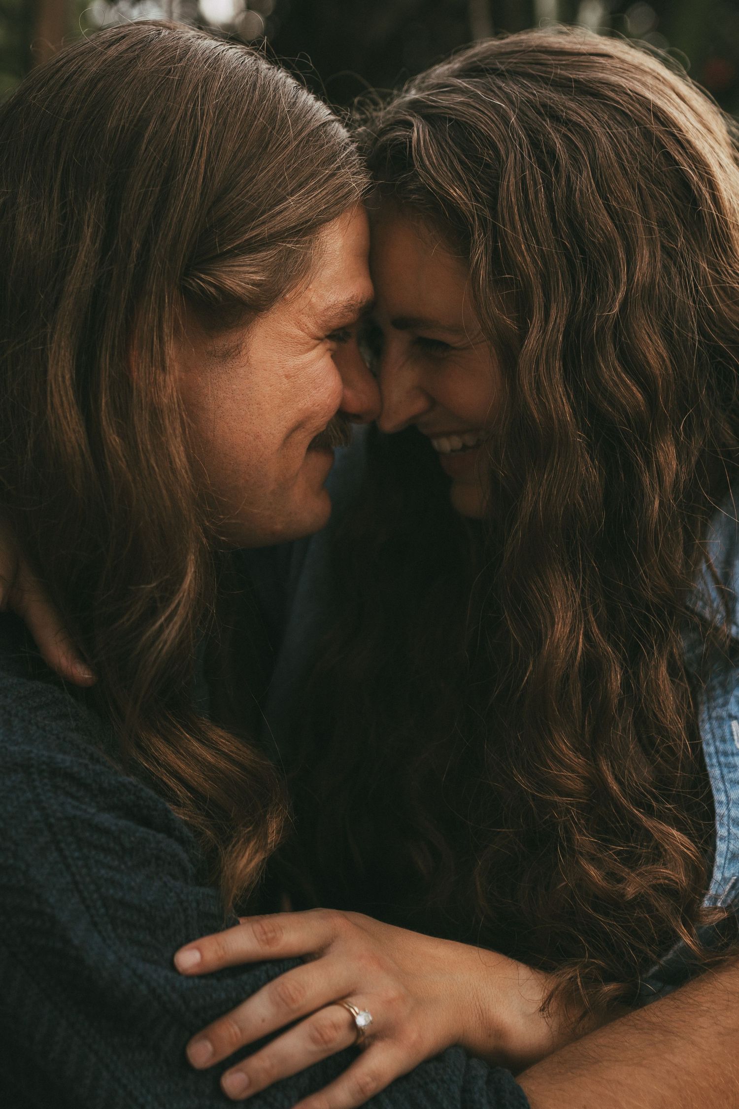 Close-up of a couple with long brown hair touching foreheads tenderly, woman wearing an engagement ring.