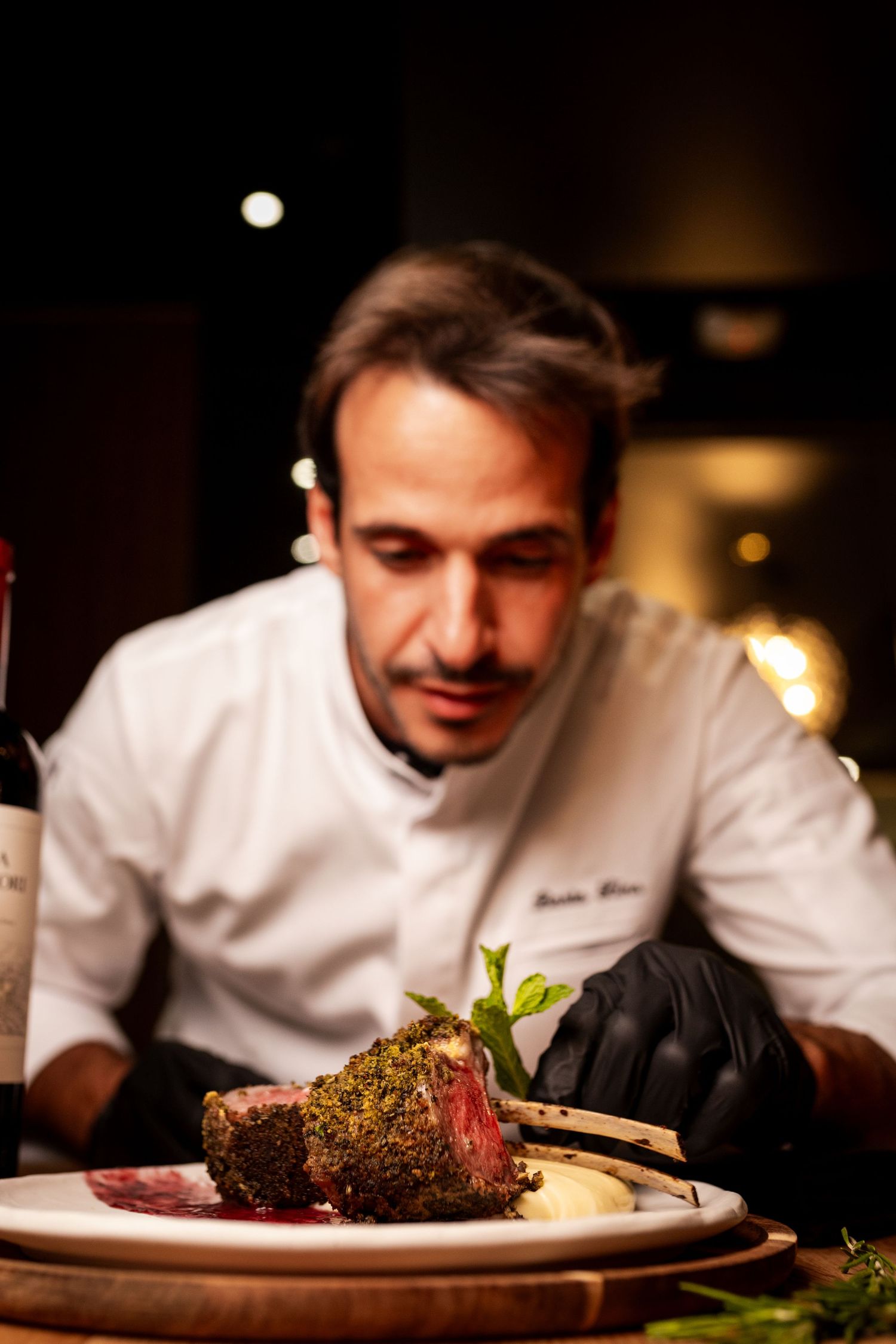 Chef Barto Lins in white uniform inspects a herb-crusted rack of lamb on a white plate in a restaurant.