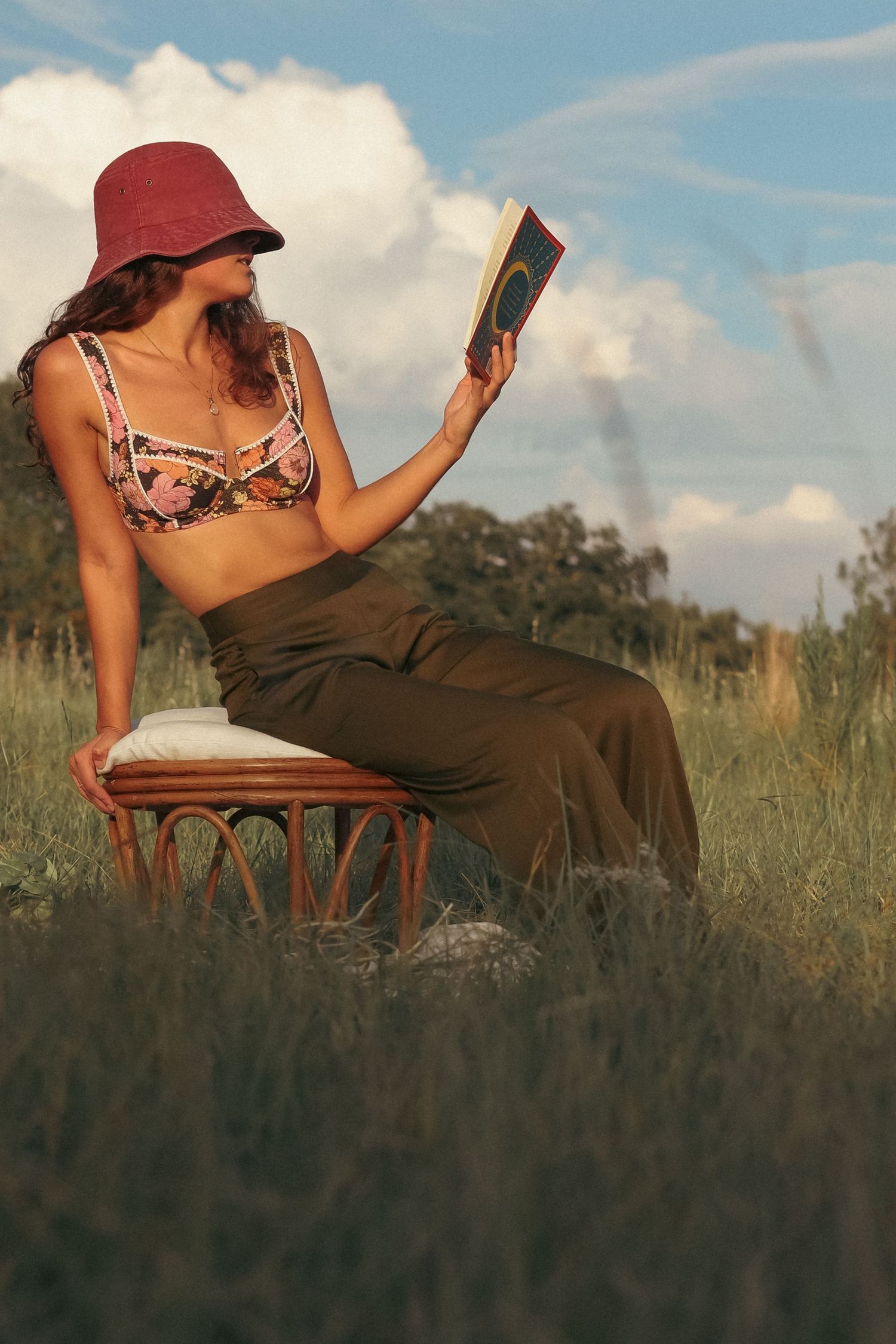 Woman in floral crop top, olive skirt, and red bucket hat reading a book outdoors in a grassy field.