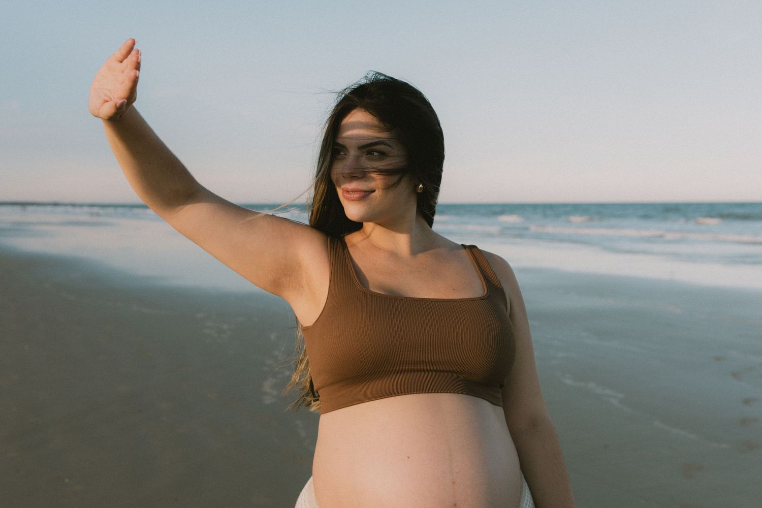 Smiling pregnant woman in brown bikini top posing on beach at sunset with waves behind her.