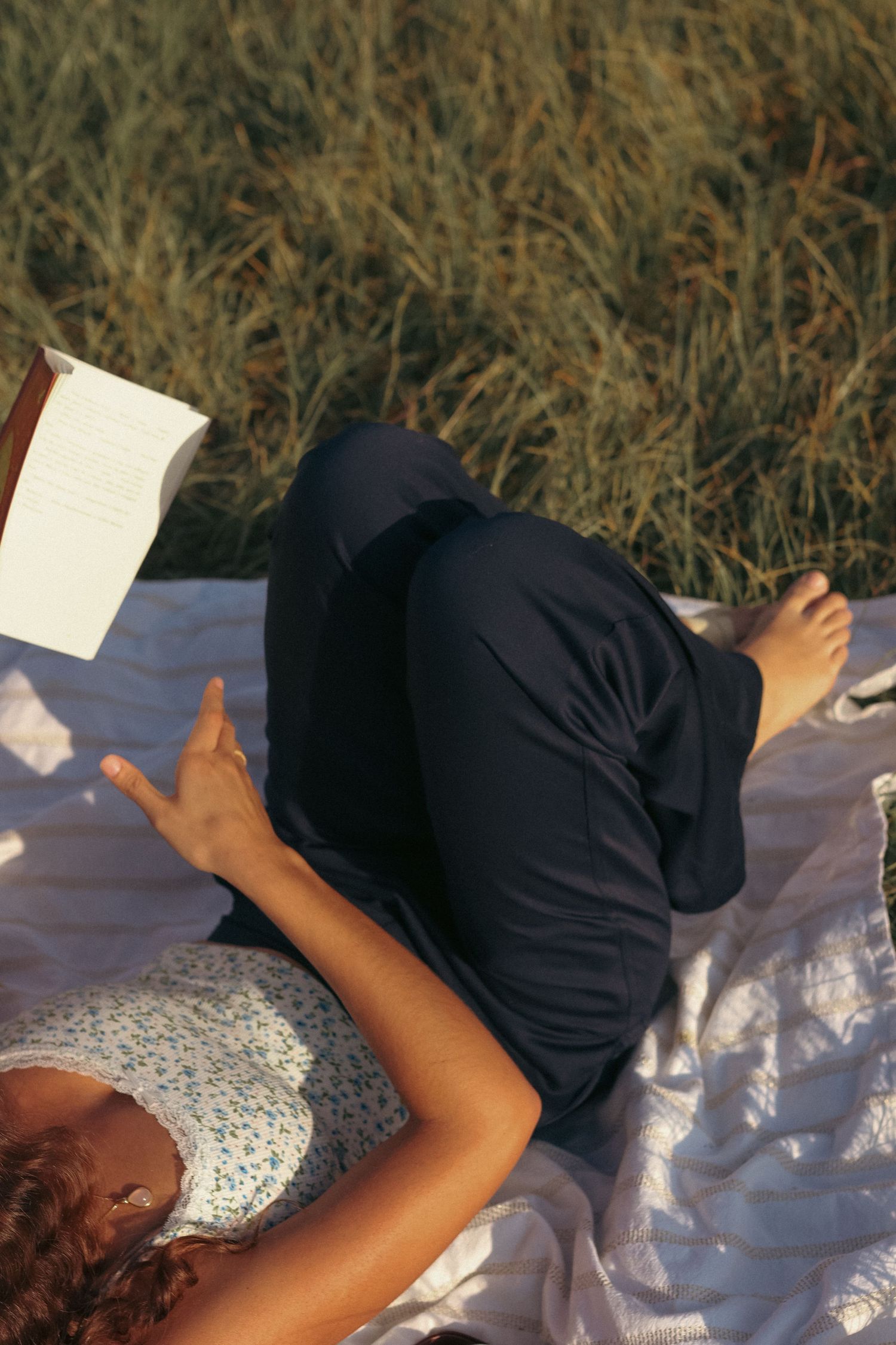 Woman lying on blanket in grass field reading book wearing floral top and navy pants.
