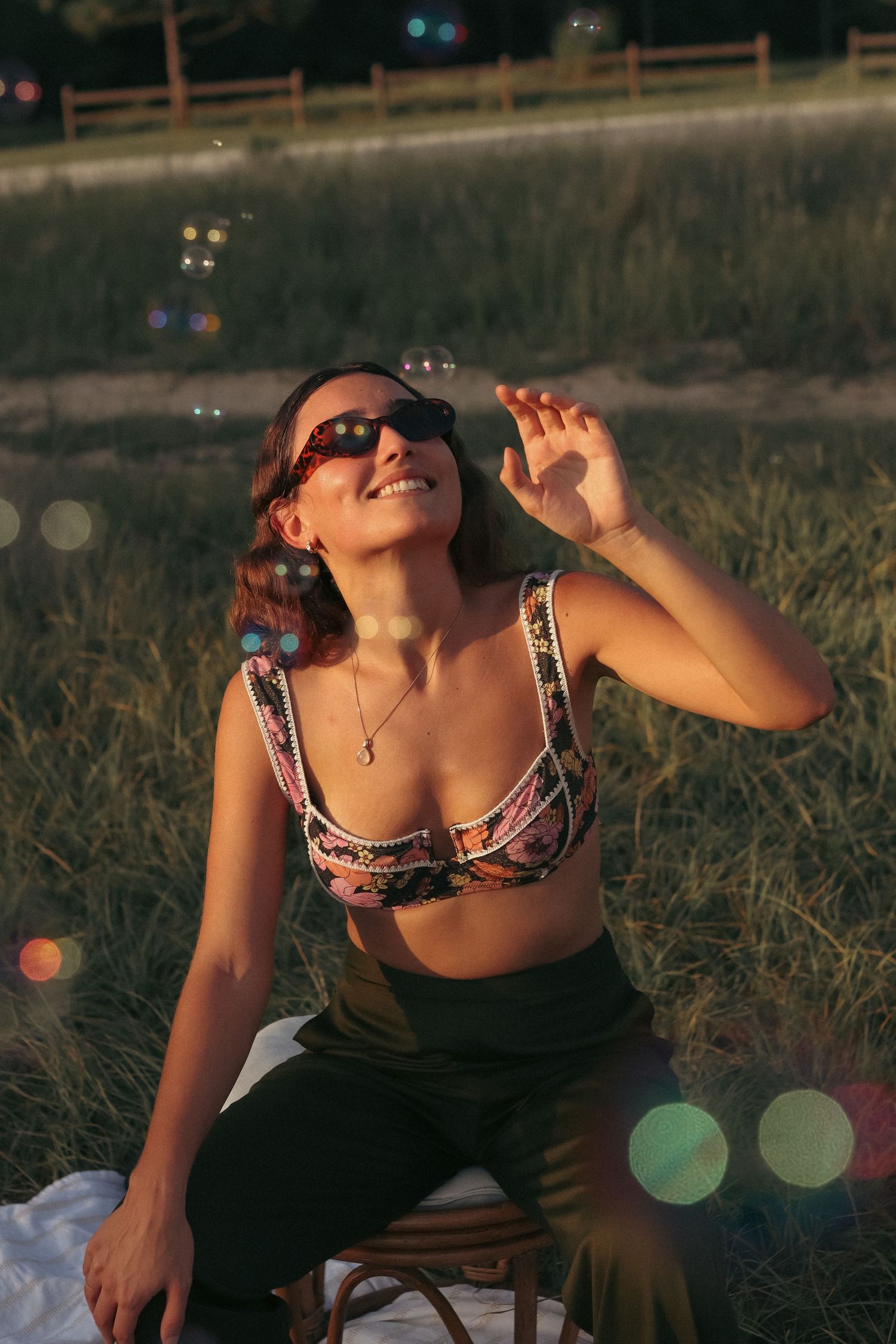 A smiling woman in sunglasses and a colorful crop top sits outdoors in golden hour light, reaching toward floating soap bubbles.