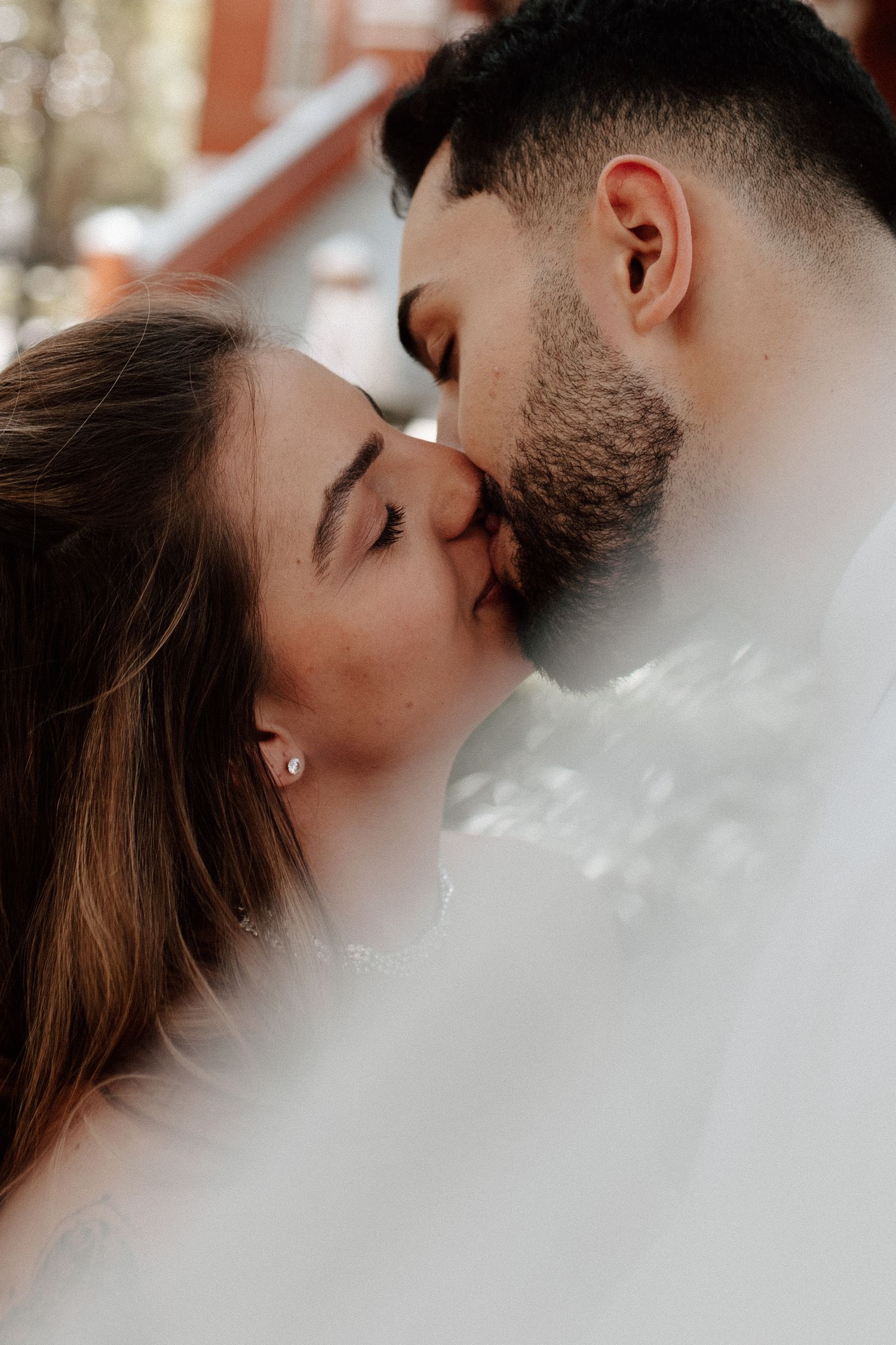 Close-up of couple sharing a kiss beneath white wedding veil in soft romantic outdoor portrait.