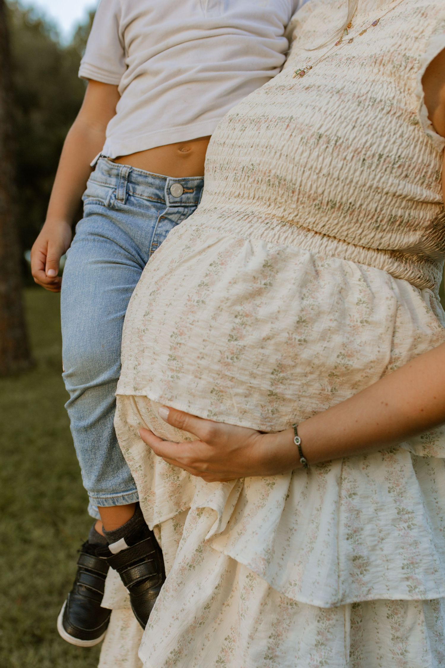 Pregnant woman in floral dress holding her baby bump beside a child in jeans outdoors.