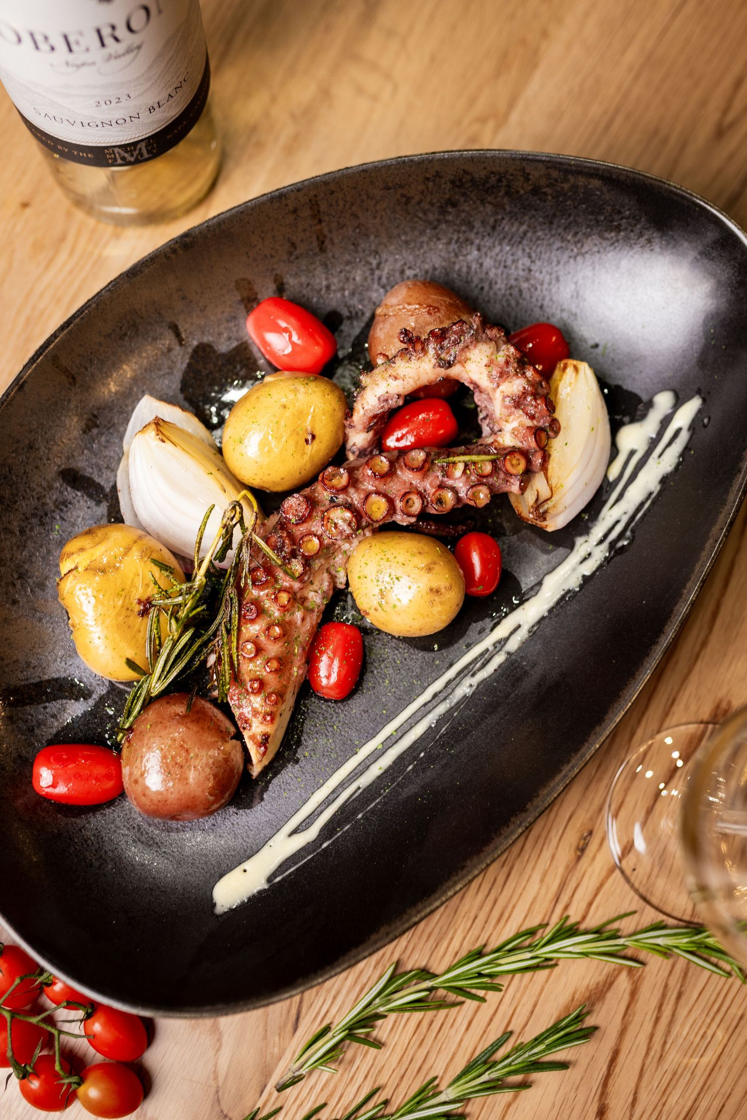Grilled octopus with cherry tomatoes, roasted potatoes, and aioli served in a black oval dish with white wine.