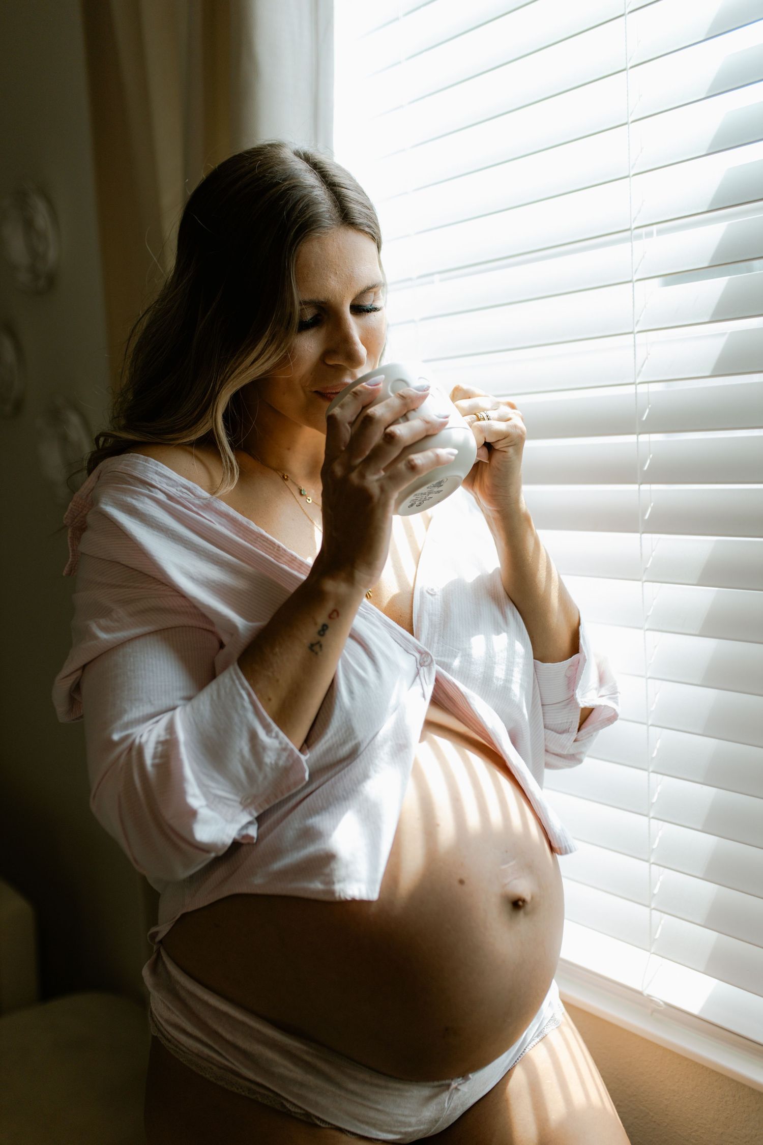 Pregnant woman in open white shirt sipping from mug while cradling bump near sunny window.