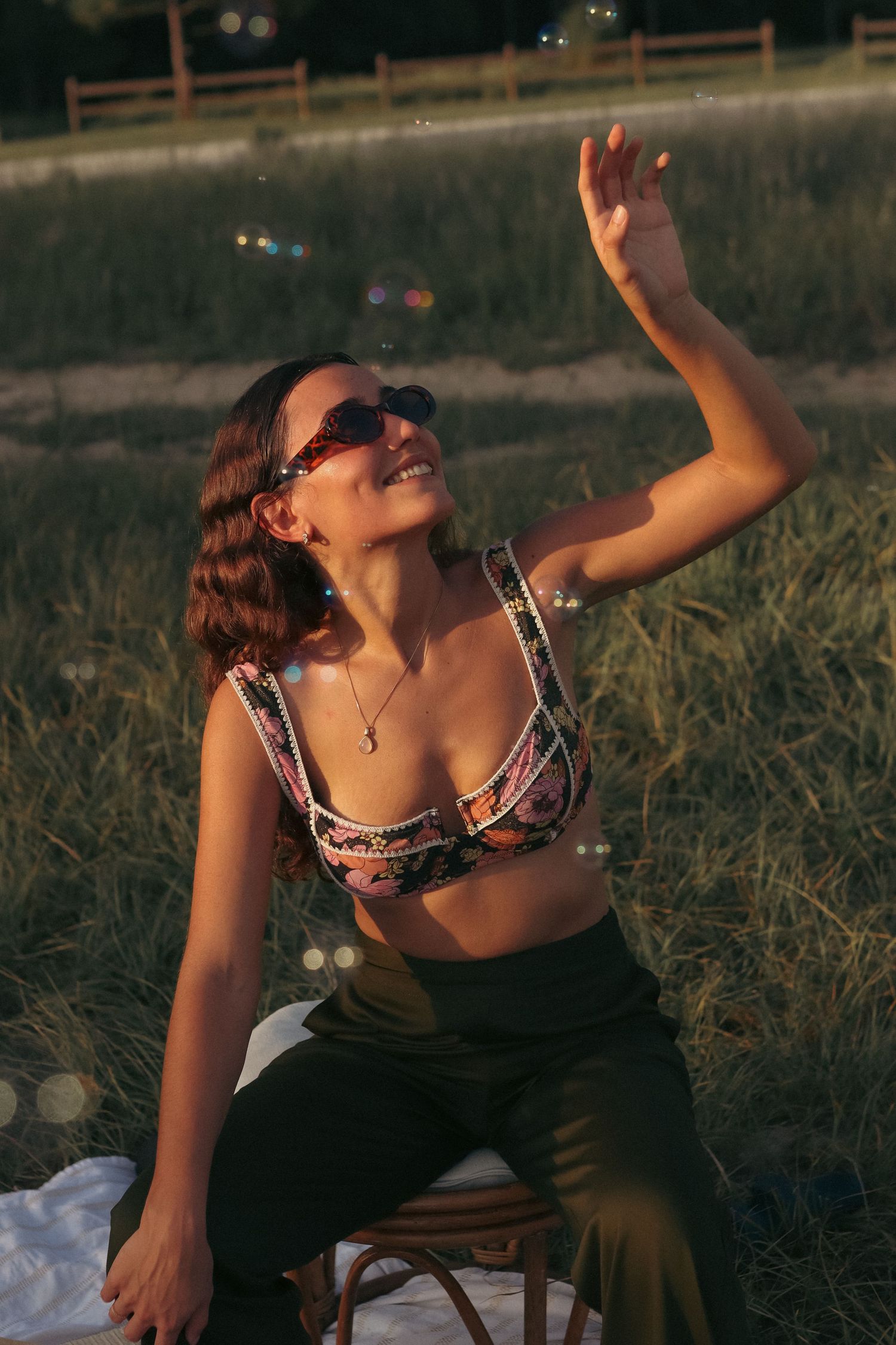 Smiling woman in embroidered crop top and sunglasses reaching for bubbles in golden sunlight.