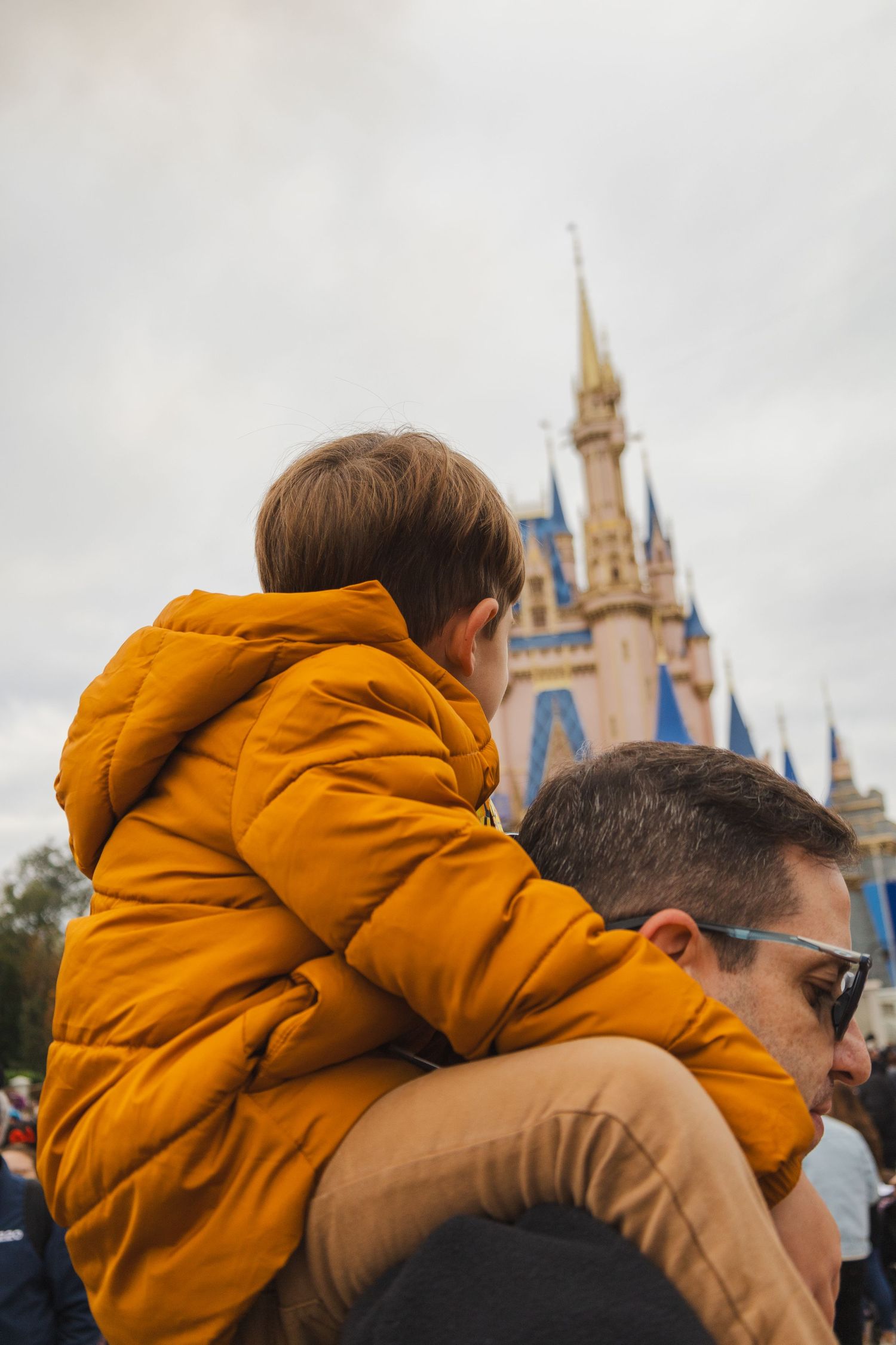 A child in a yellow jacket sits on a man's shoulders in front of Cinderella's Castle at Walt Disney World.