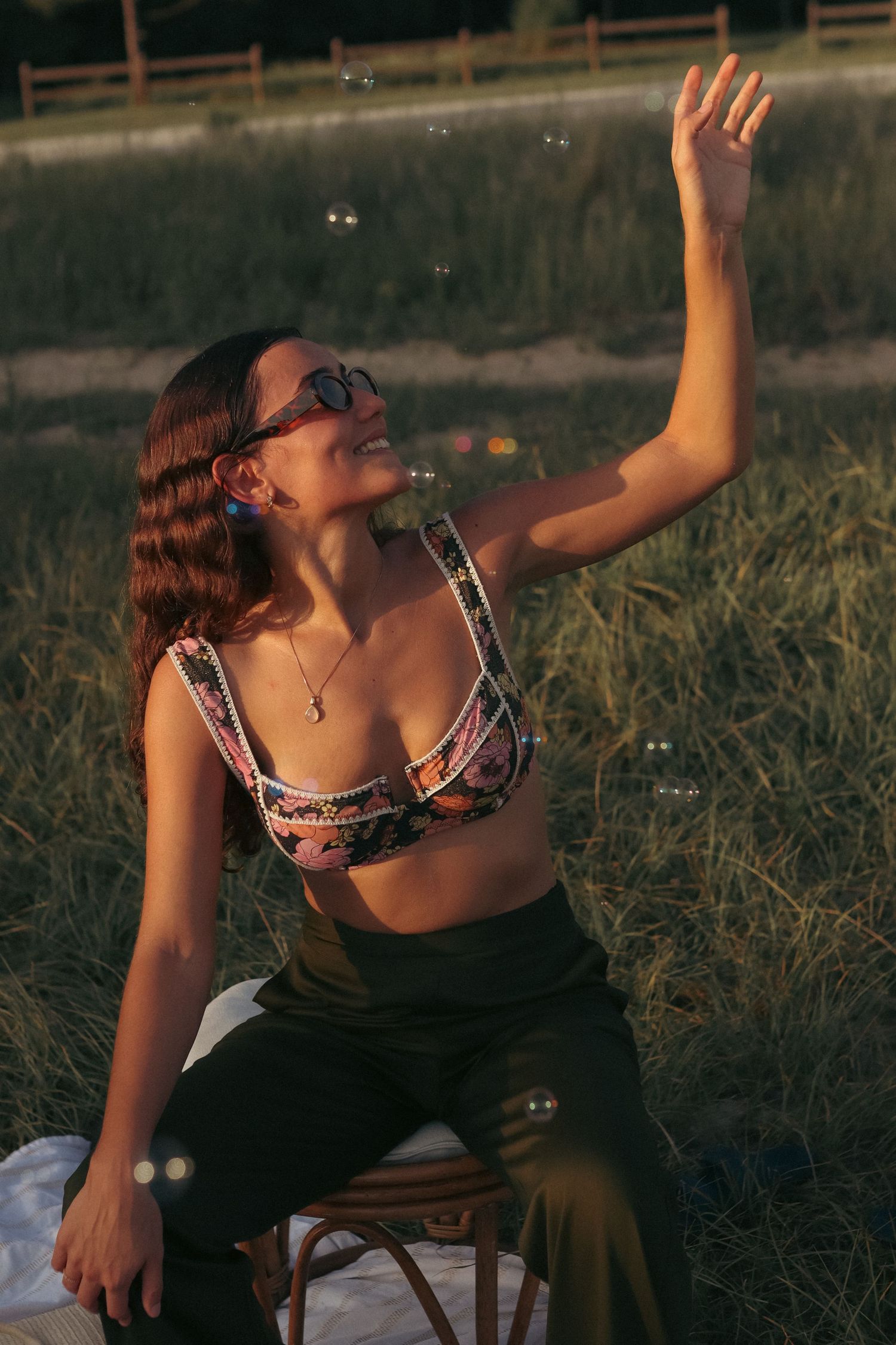 Young woman in crop top and sunglasses reaching for soap bubbles outdoors in golden hour light.