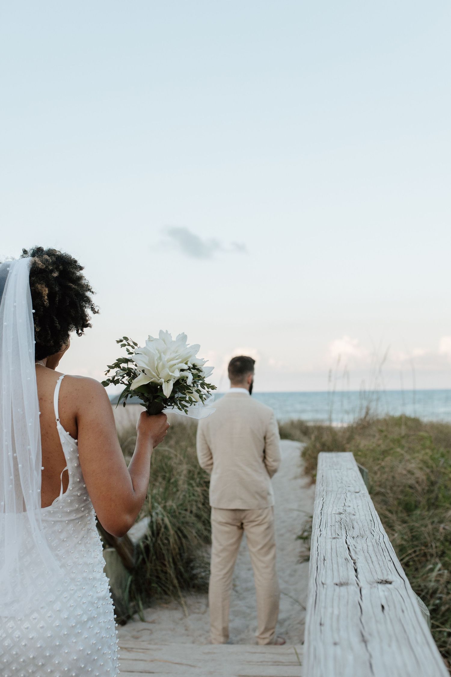 A bride in a white beaded gown holding a white lily bouquet approaches her groom on a beach boardwalk.