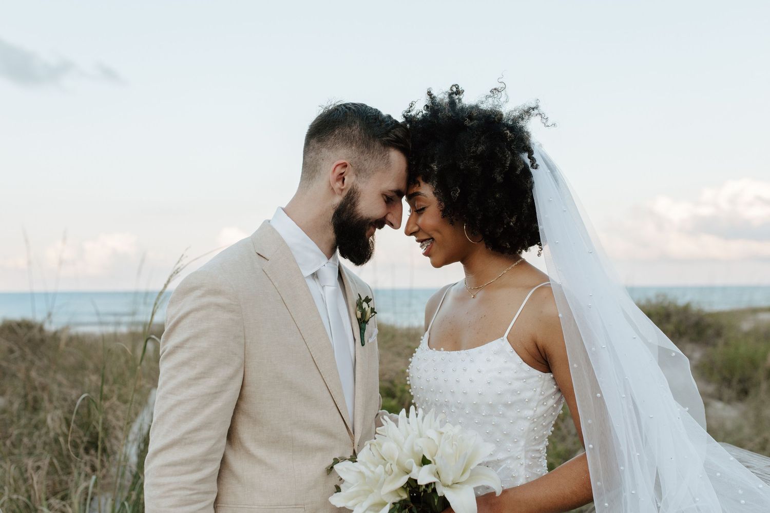 Interracial bride and groom touching foreheads romantically on beach, bride holding white bouquet at sunset.