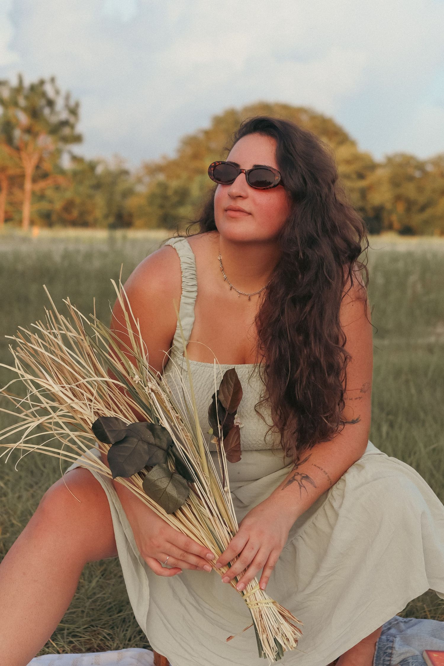 Curly-haired woman in sage sundress and sunglasses holding dried wheat bouquet in a field.