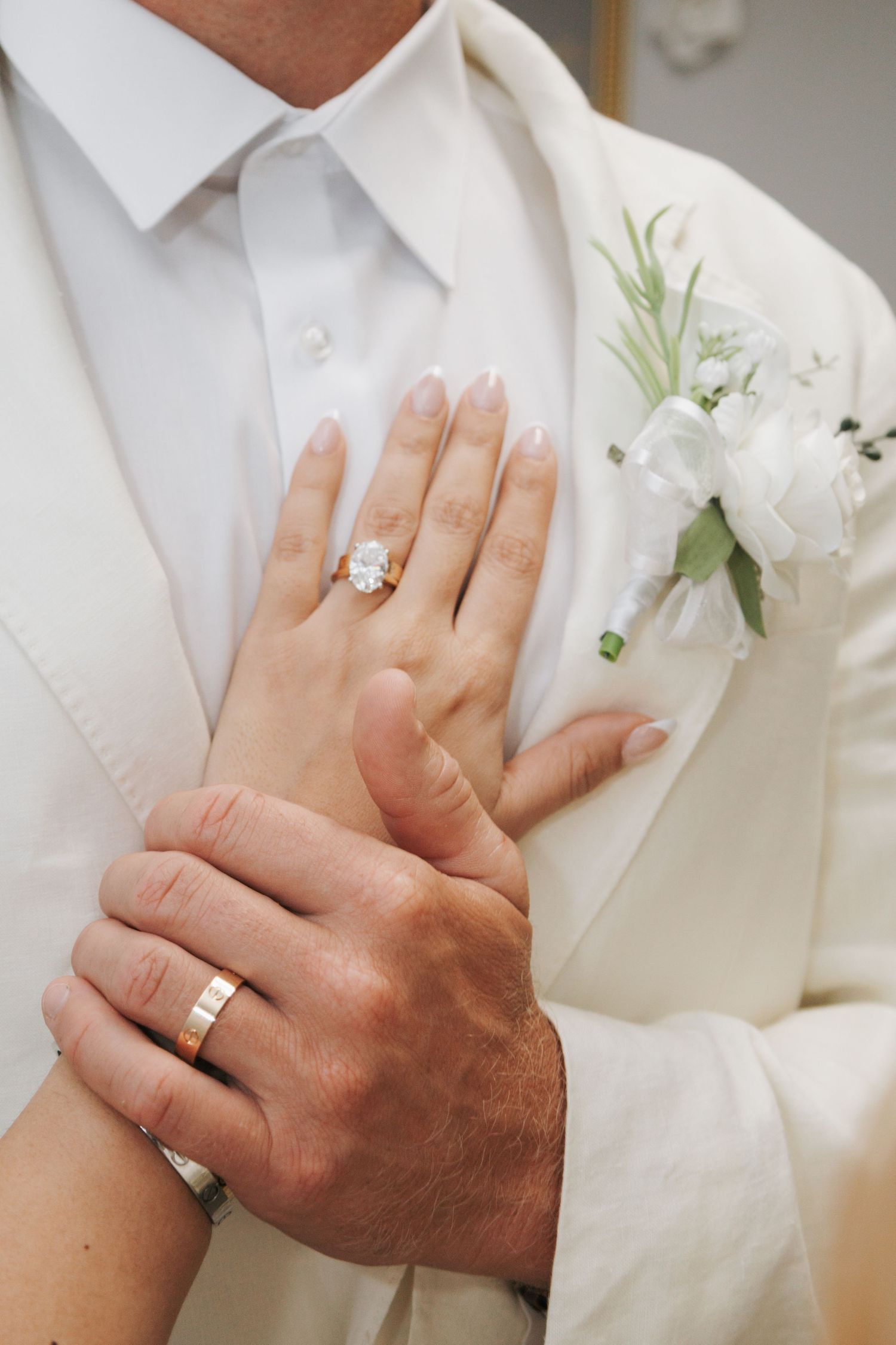 Bride's hand with diamond engagement ring resting on groom's chest in a white suit with boutonniere.