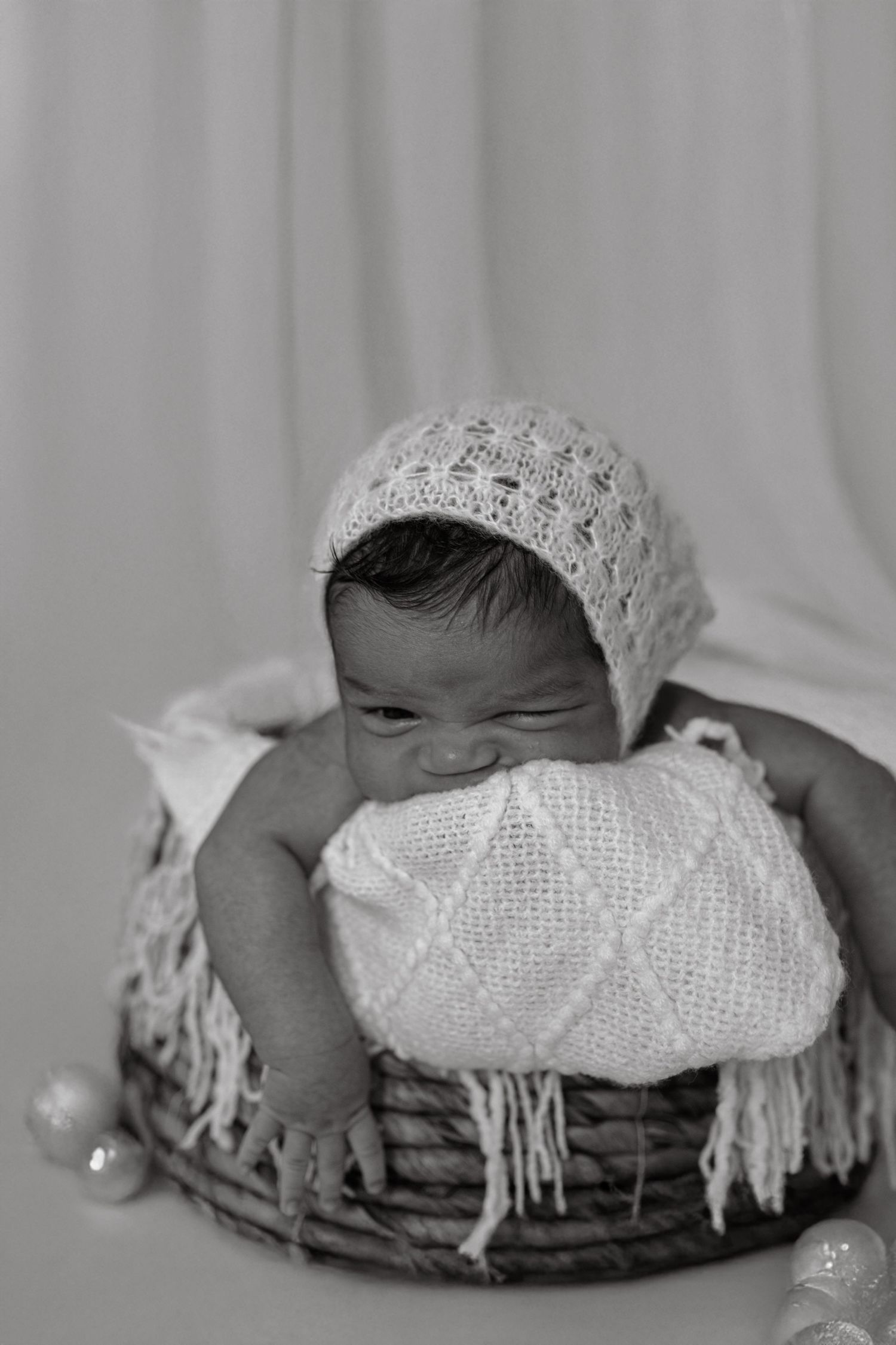 Adorable newborn baby in knit bonnet sitting in wicker basket with white blanket.