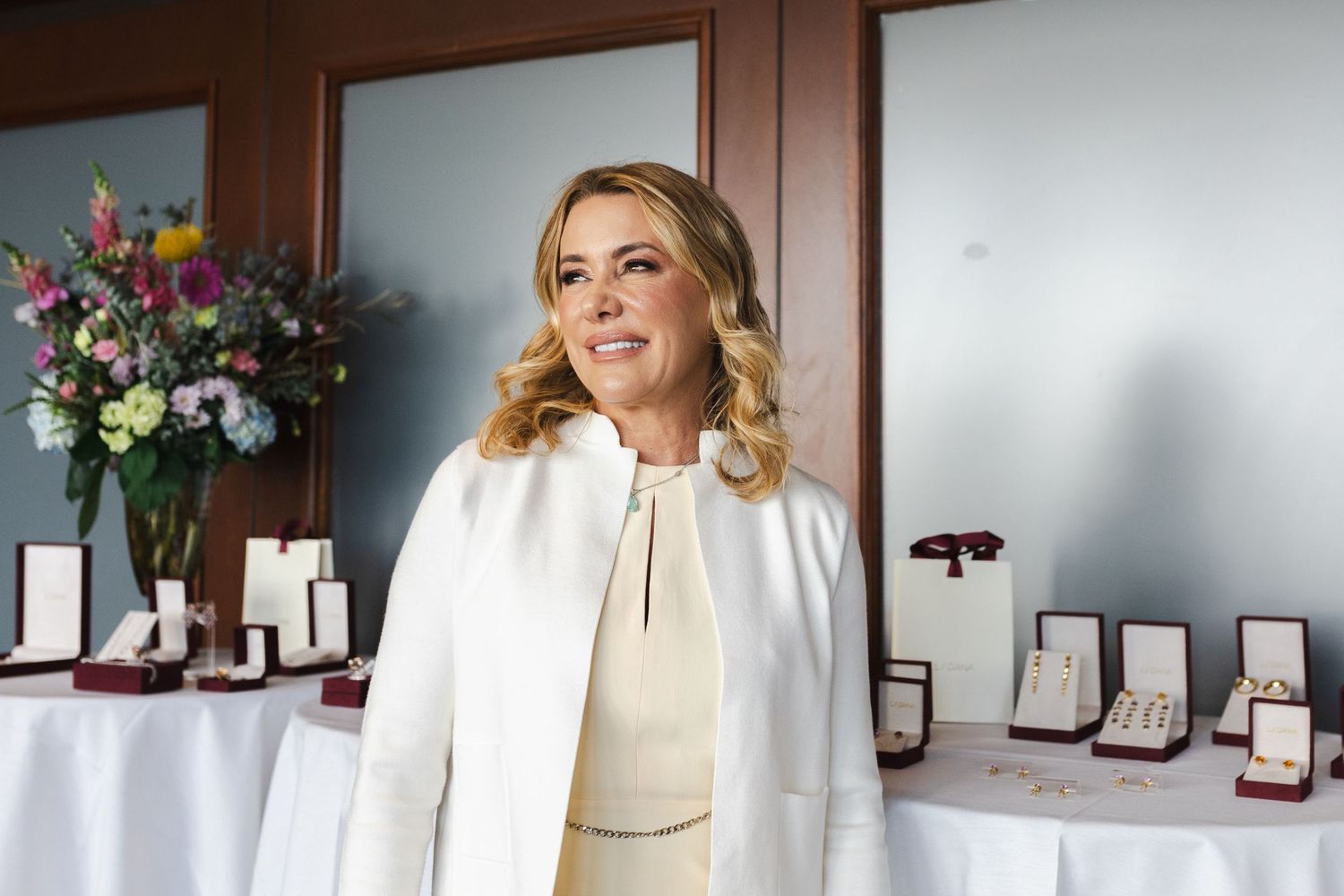 Blonde woman in white blazer standing before a jewelry display table with floral arrangement in background.