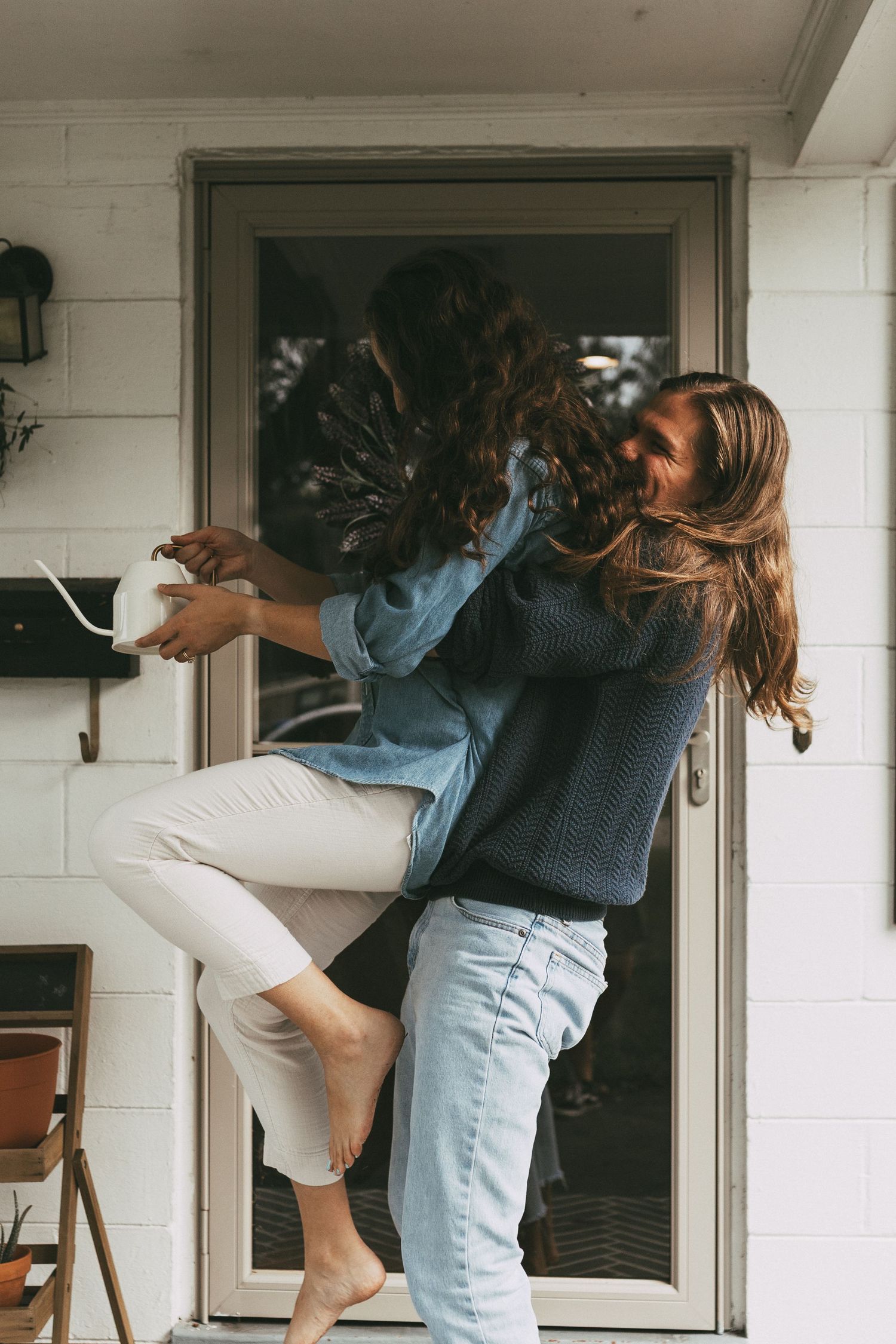 A playful couple embraces and spins on a white brick porch, woman barefoot in white pants and denim shirt.