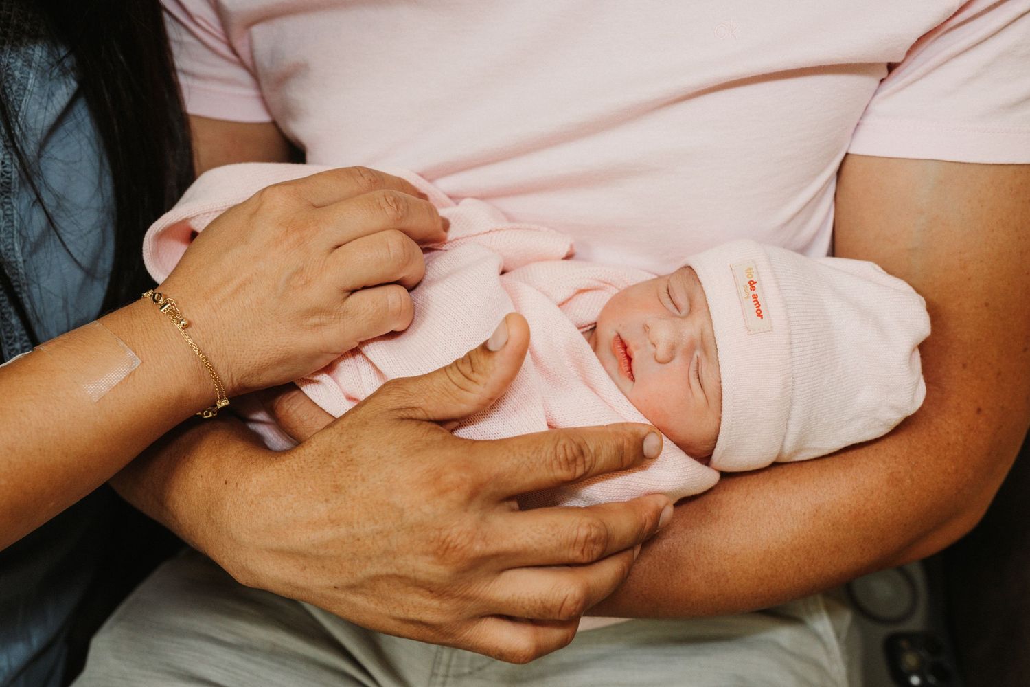 Newborn baby swaddled in pink blanket and hat, cradled in parents arms.