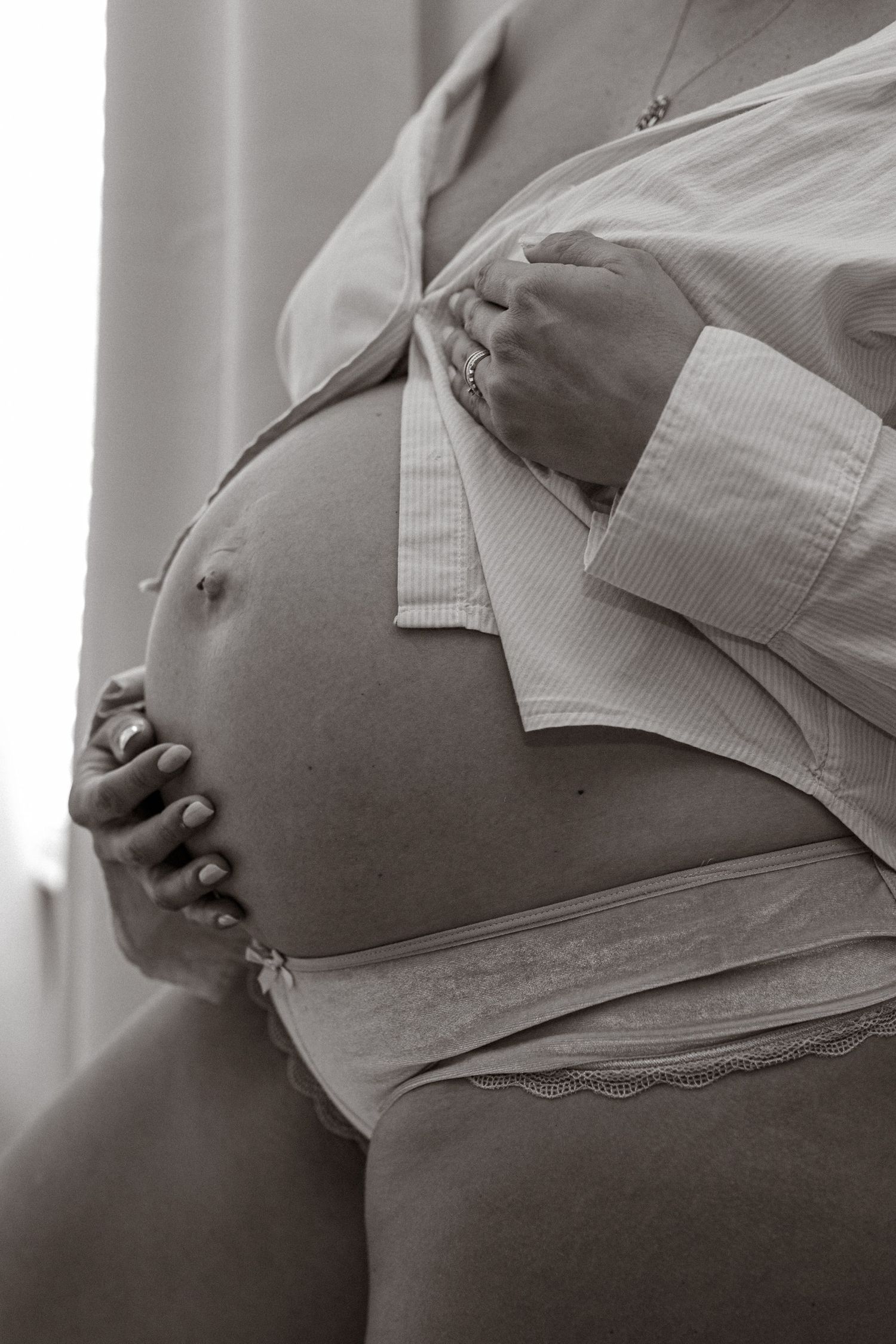 Black and white close-up of pregnant woman in open white shirt and lace underwear holding bump.