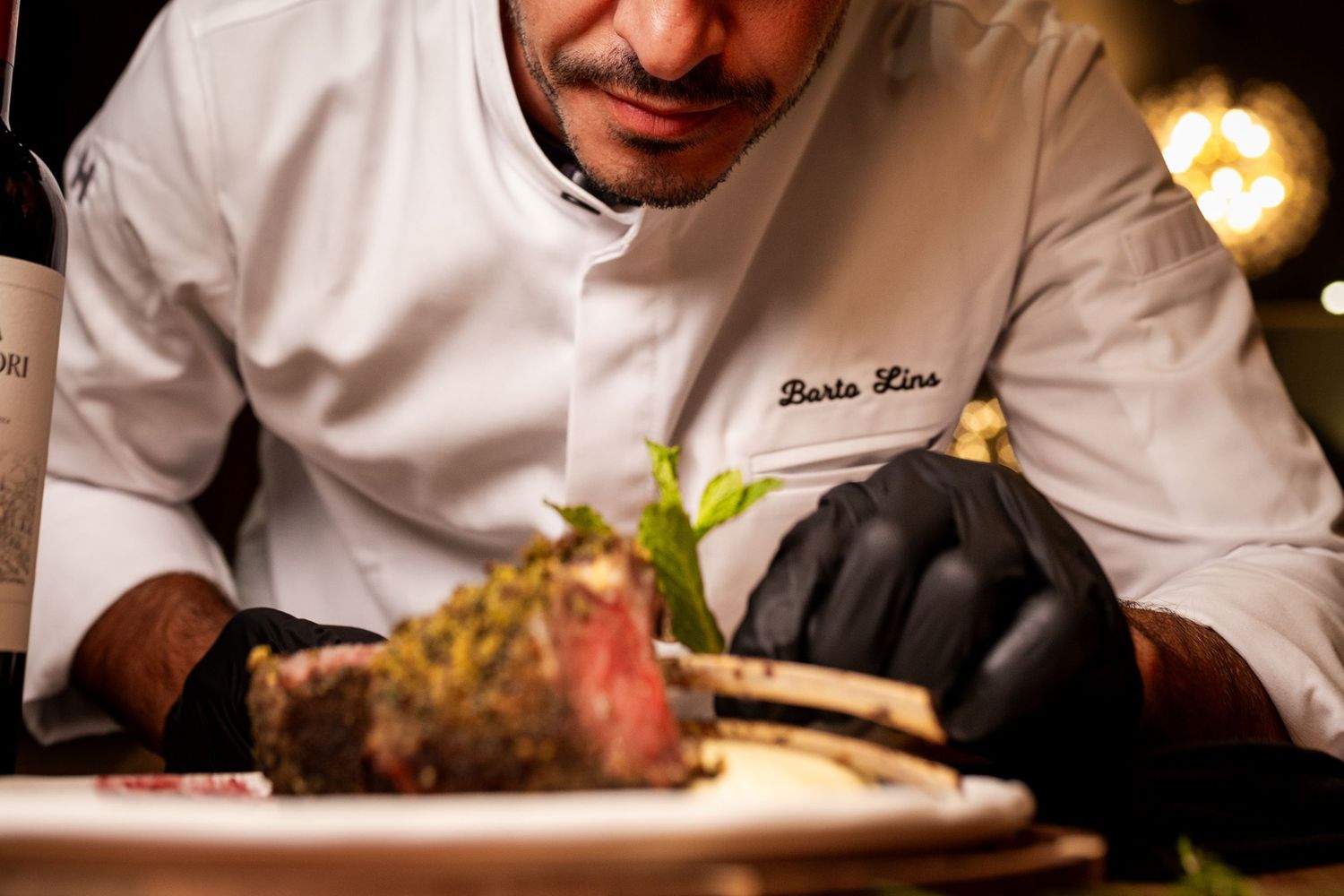 Chef Barto Lins in white jacket and black gloves carefully garnishes a herb-crusted rack of lamb on a plate.